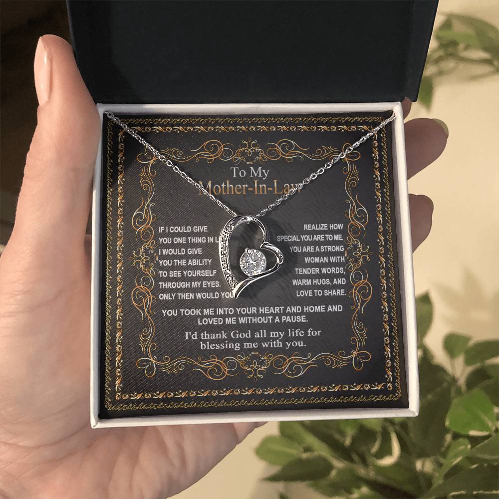 A person holds a "To Mother-In-Law, Through My Eyes - Forever Love Necklace" with a heart-shaped pendant in a gift box. The box is open, revealing a heartfelt message dedicated to a mother-in-law, making it the perfect gift for your mom.