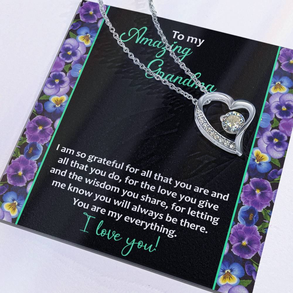 A Forever Love Necklace with a heart-shaped pendant adorned with a cubic zirconia stone rests on a card decorated with purple flowers and a heartfelt message. The shimmering 14k white gold finish highlights the text, "To Grandma, My Everything" and "I love you!