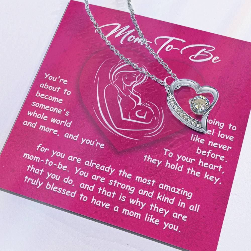 The "To Mom To Be, Someone's Whole World - Forever Love Necklace" features a heart-shaped pendant with a central gemstone, elegantly displayed on a card. The card is thoughtfully designed for moms-to-be, adorned with a heartfelt message set against a pink background and an illustration of a pregnant woman. This personalized gift beautifully captures the essence of everlasting love and anticipation.