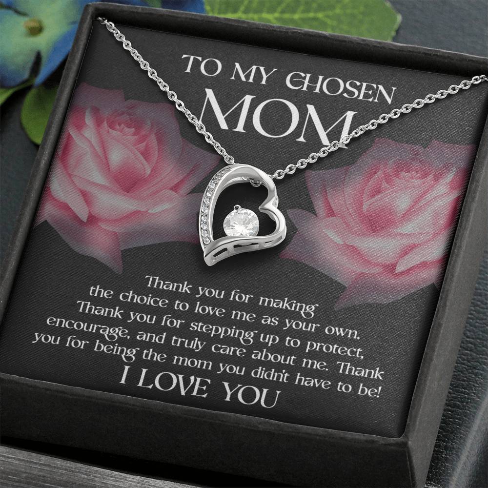A beautiful heart-shaped pendant necklace with a gem is displayed in an open box. The "To Bonus Mom, My Chosen Mom - Forever Love Necklace" boasts a stunning yellow gold finish and features a heartfelt message addressed “To My Chosen Mom” set against a backdrop of pink roses, making it the ideal Mother’s Day gift.