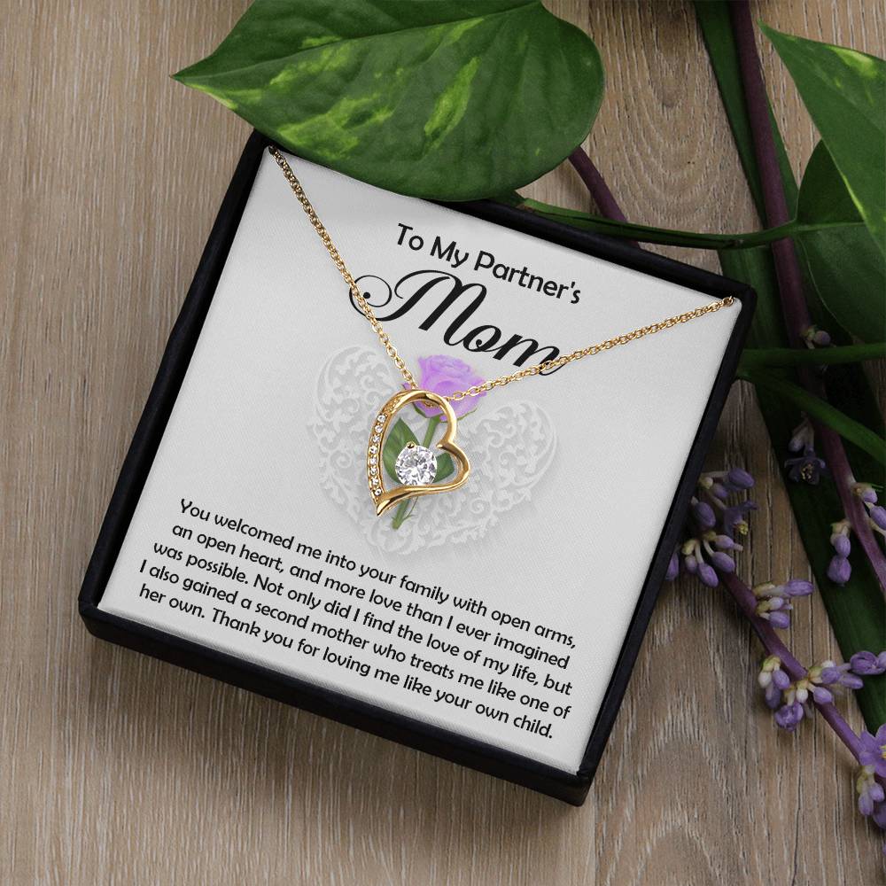 The Partner's Mom-A Second Mother - Forever Love Necklace, gold-finished with clear cubic zirconia, is shown in an open black gift box. Inside, a heartfelt message for a partner’s mom sits on wood with green leaves and purple flowers.
