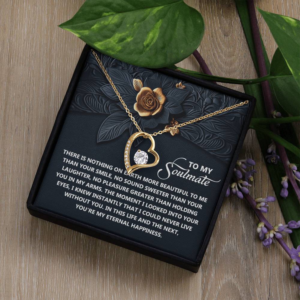 The Soulmate-Eternal Happiness - Forever Love Necklace, a gold heart-shaped piece with a dazzling CZ crystal, rests in an open jewelry box. Its lid features an embossed floral design and a heartfelt love and happiness message, elegantly set against a wood surface with scattered leaves and flowers.