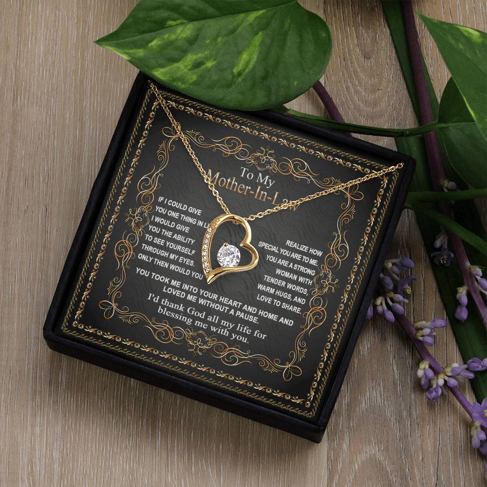 A "To Mother-In-Law, Through My Eyes - Forever Love Necklace" with a heart-shaped pendant is placed on a decorative box containing a heartfelt message addressed to a Mother-In-Law, surrounded by green leaves and purple flowers.