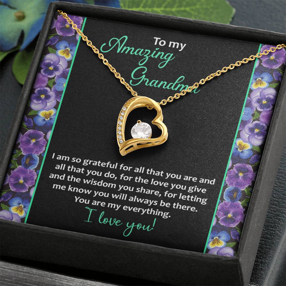 The "To Grandma, My Everything - Forever Love Necklace" features a heart pendant adorned with a stunning cubic zirconia stone and is beautifully presented in a box. The lid bears a heartfelt message addressed to "Grandma," expressing gratitude and love. The background is elegantly decorated with purple flowers, enhancing the charm of this 14k white gold finish piece.
