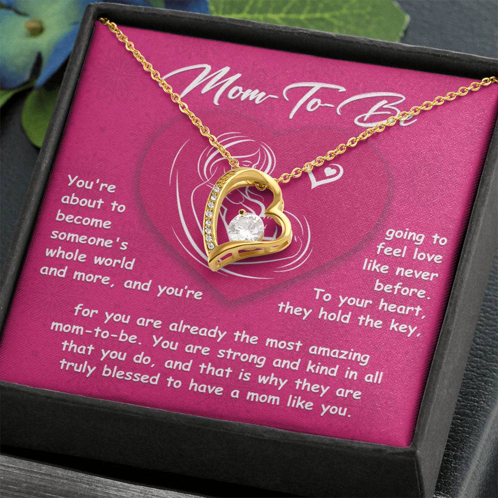 A gold necklace with a heart-shaped pendant containing a gem is showcased in an elegant jewelry box set against a pink background, featuring the inscription "To Mom To Be, Someone's Whole World - Forever Love Necklace.
