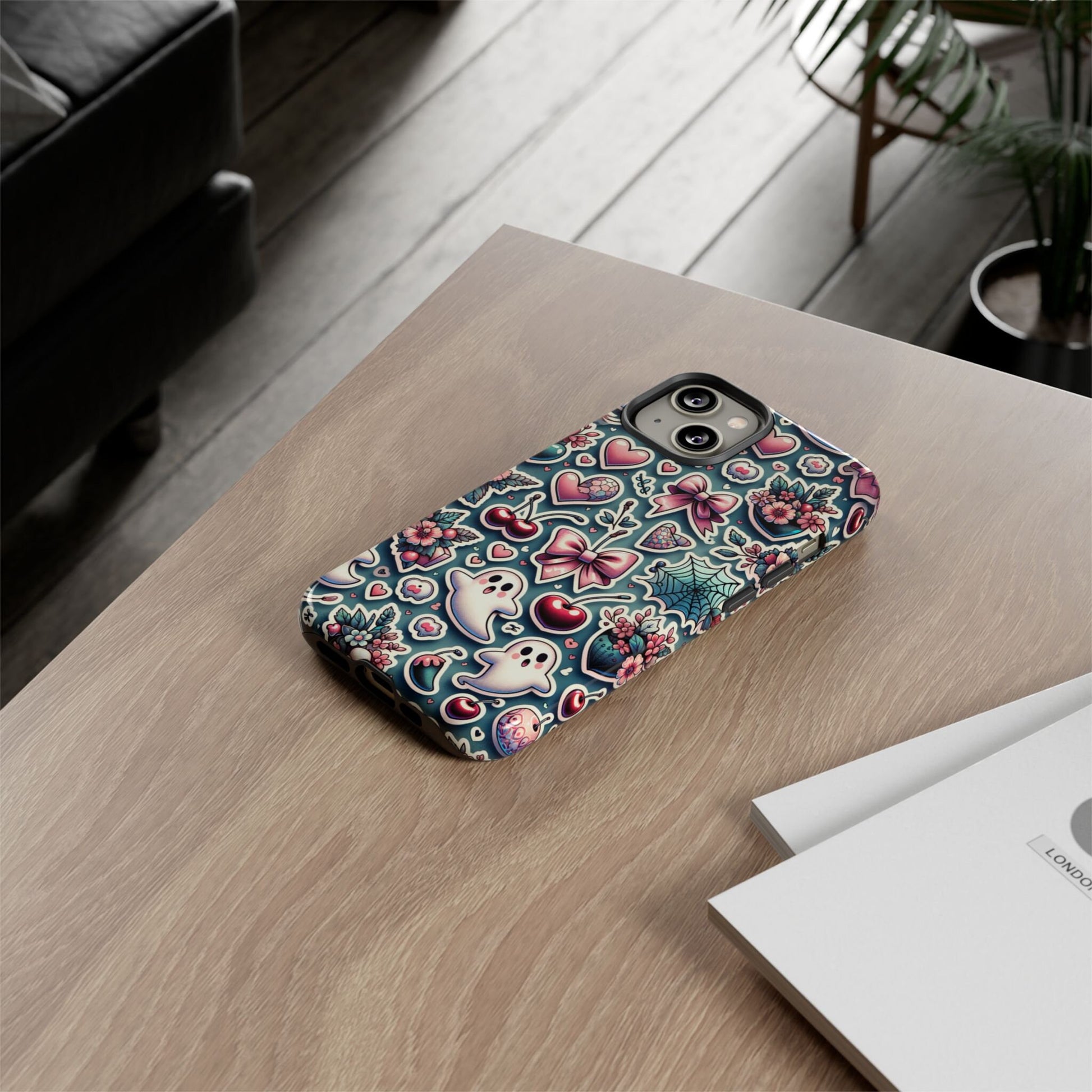 A smartphone with a Cute Coquette Halloween Phone Case, adorned with hearts and floral designs and embodying the soft girl aesthetic, rests on a wooden table next to a book and a potted plant.