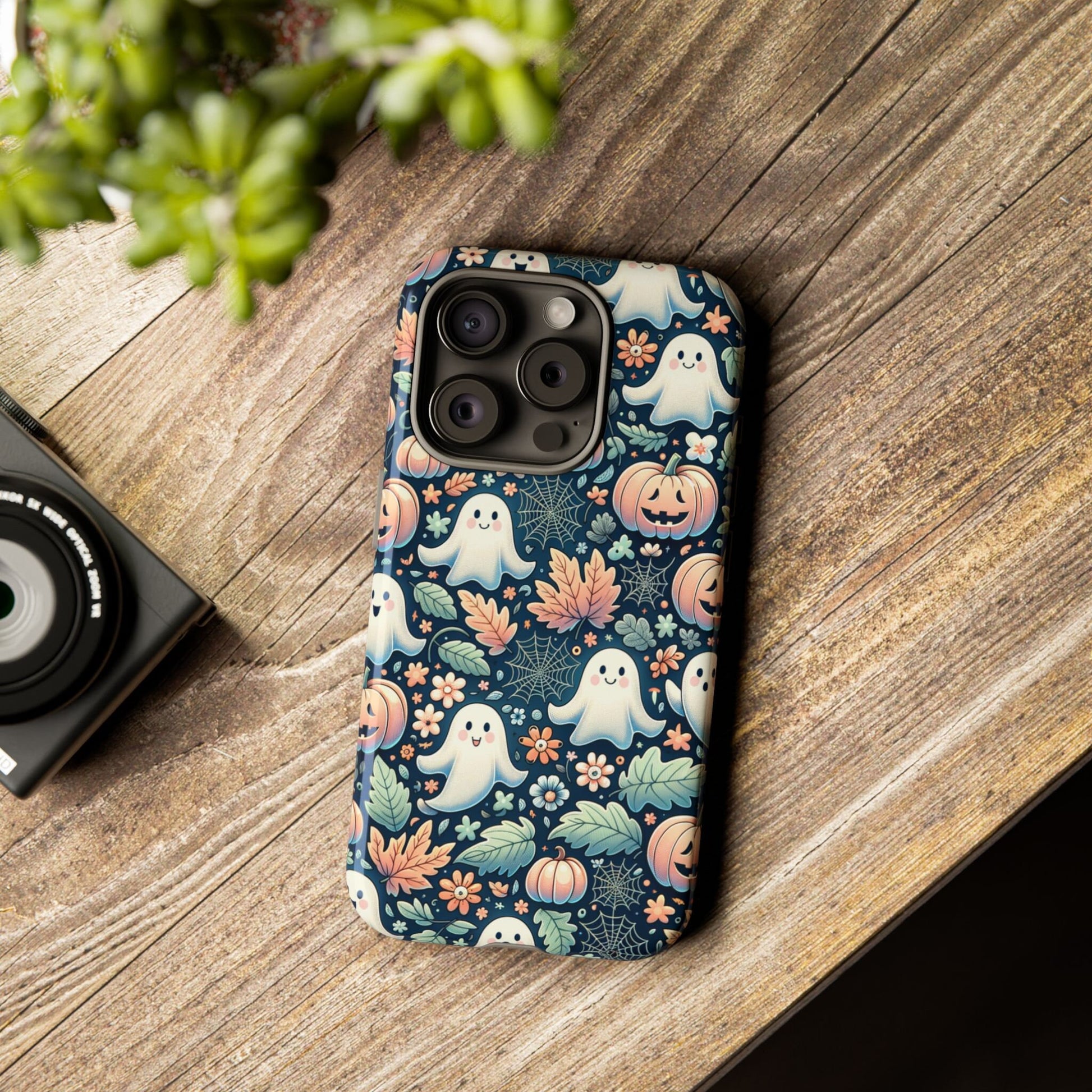 The "Happy Halloween Cute Ghost And Pumpkin Phone Case," perfect for iPhone 15, 14, 13 Pro, Samsung Galaxy, and Pixel users who are Halloween lovers, features a delightful pattern of ghosts and pumpkins. It rests on a wooden surface beside a camera and a plant.