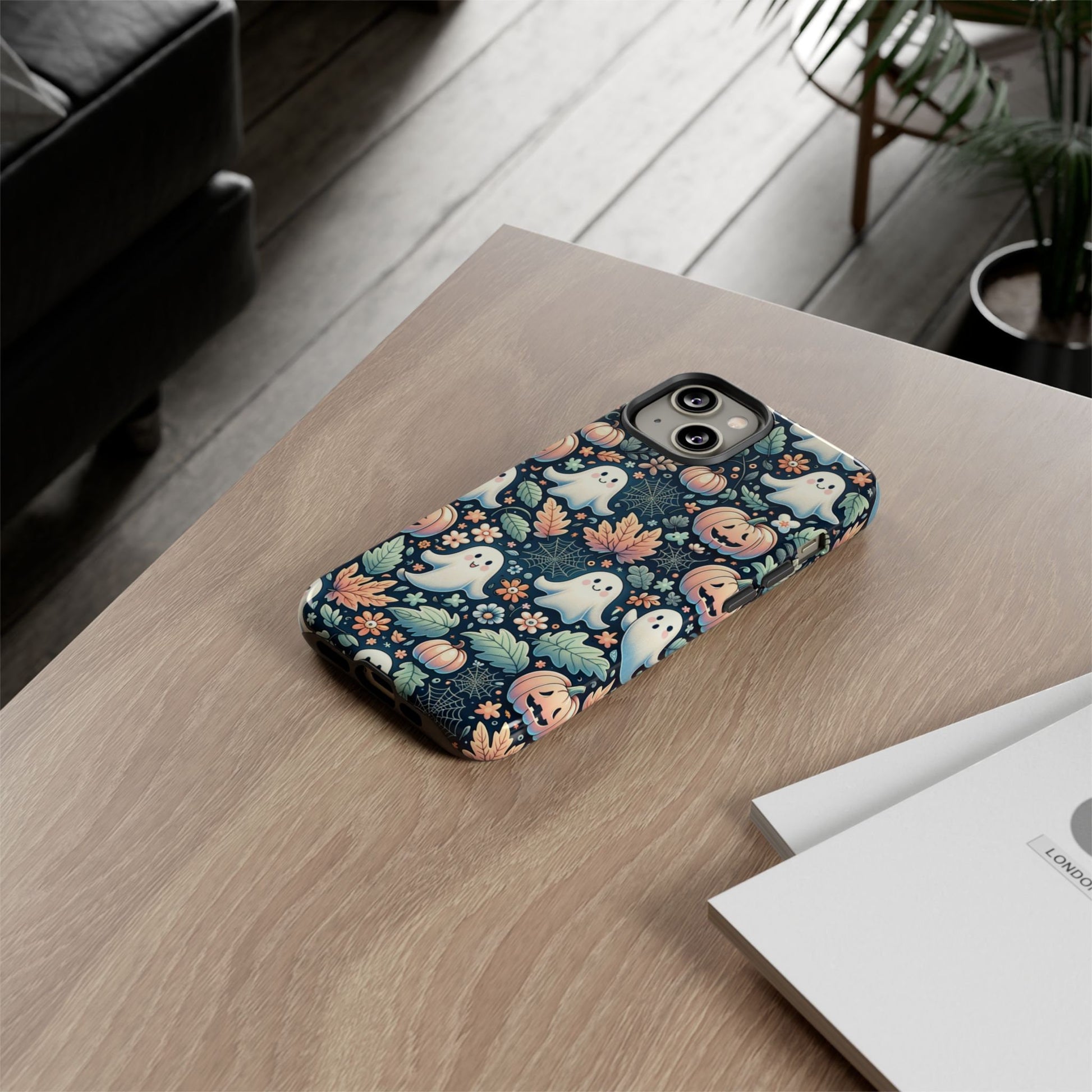 An iPhone with the Happy Halloween Cute Ghost And Pumpkin Phone Case, a perfect gift for Halloween lovers during the spooky season, sits on a wooden table next to a stack of papers.