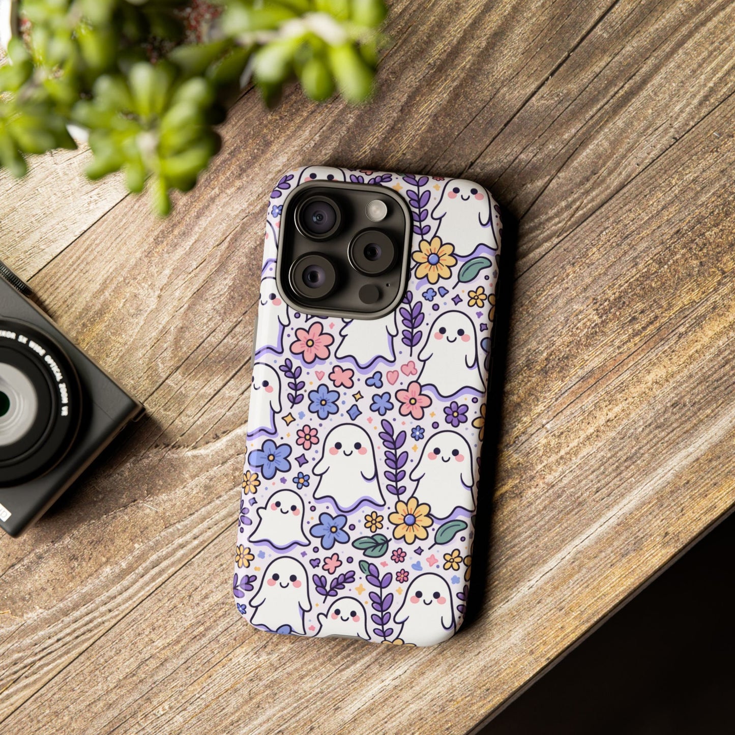 A smartphone with a Cute Ghosts And Flowers Phone Case, featuring charming cartoon ghosts and flowers, sits on a wooden table next to a small green plant and a camera.