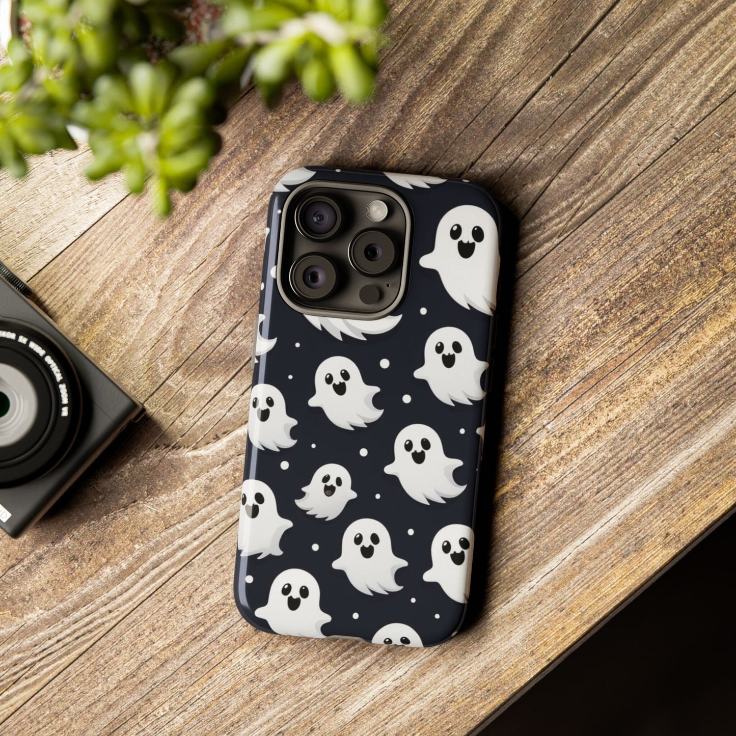 A smartphone sits on a wooden table, sporting a Cute Little Ghosts Phone Case, an ideal gift for Halloween enthusiasts. Nearby, a camera and a potted plant enhance the charm of the scene.