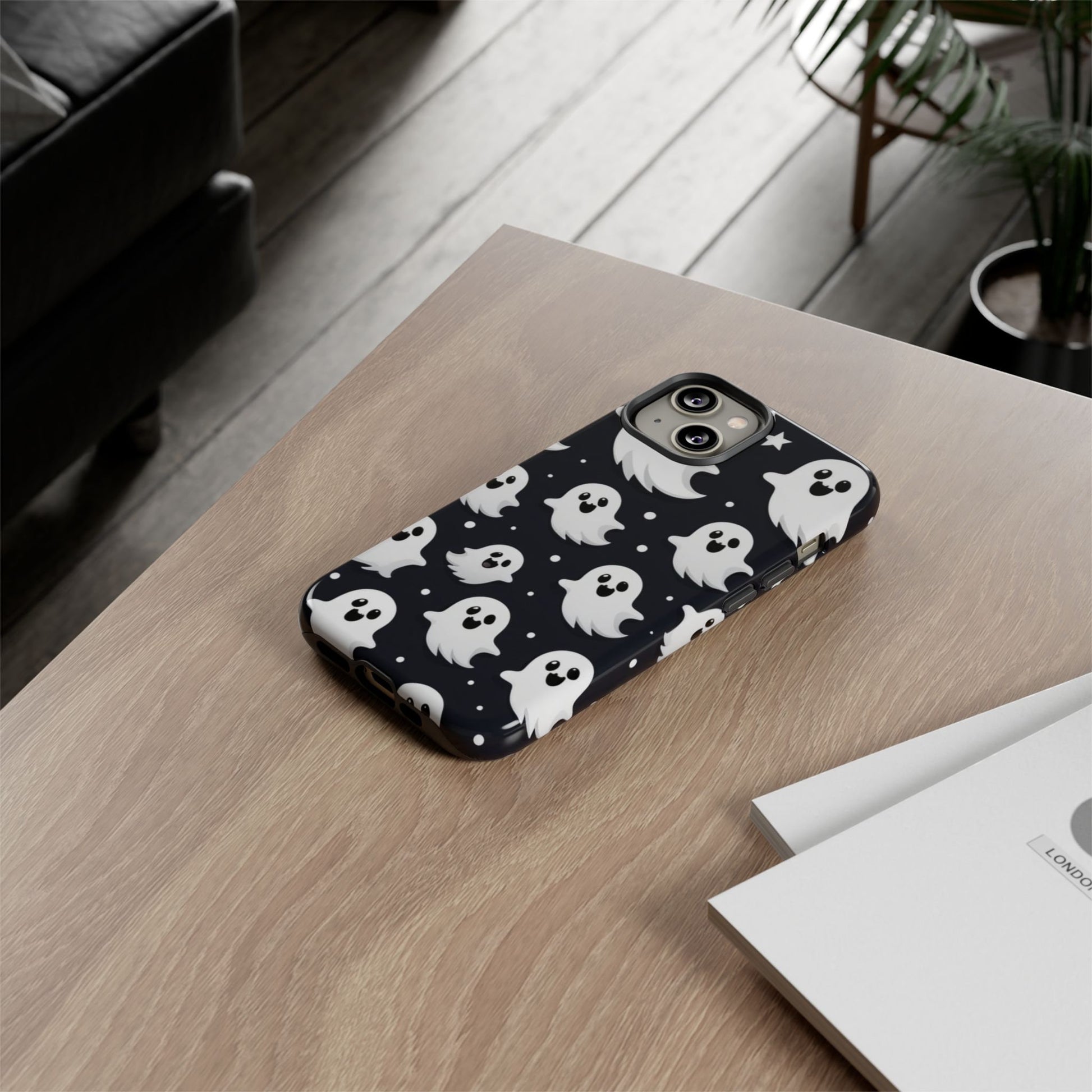 A smartphone adorned with a Creepy Cute Ghosts Case, an ideal Halloween gift for enthusiasts, rests on a wooden table beside a booklet.