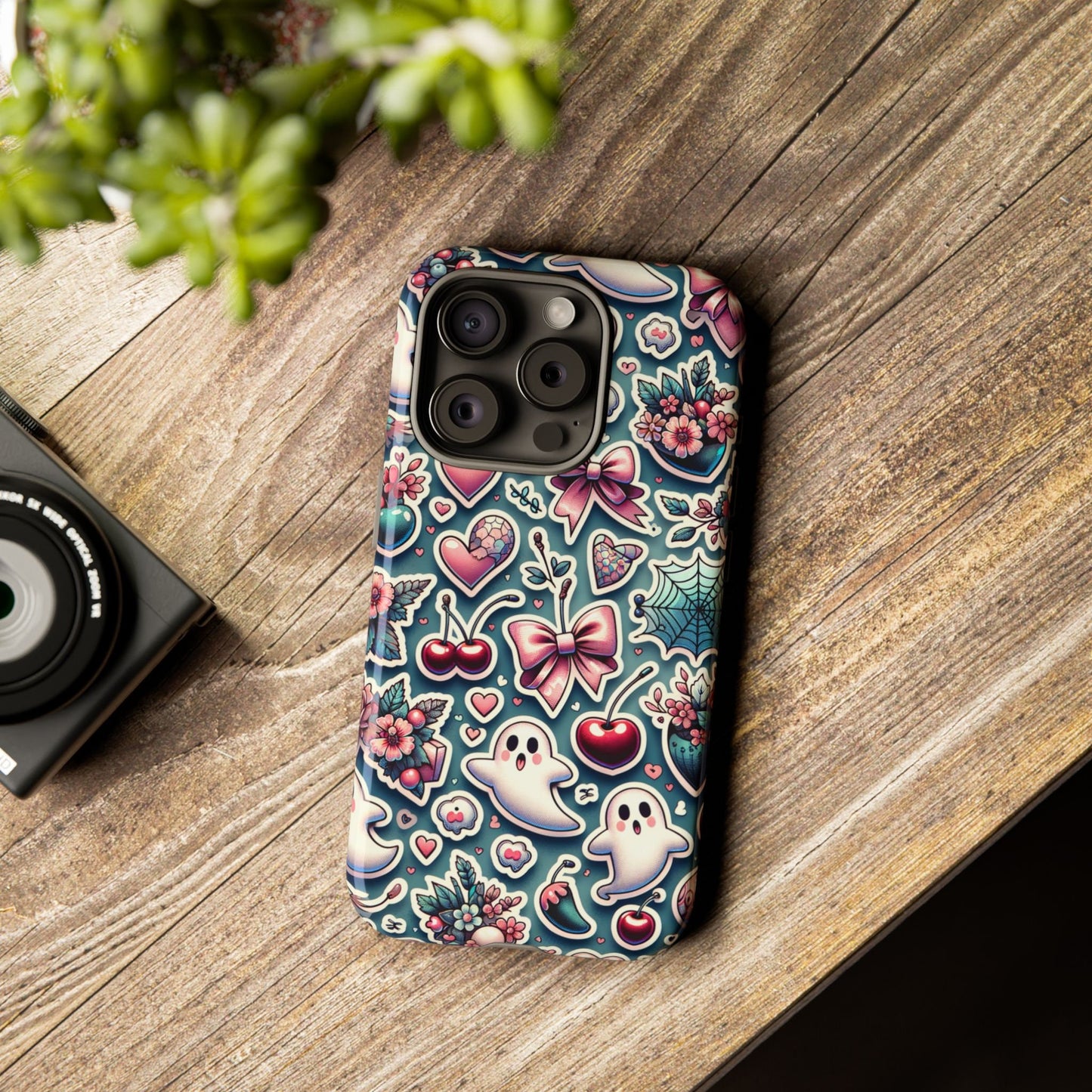 An iPhone with a Cute Coquette Halloween Phone Case featuring a coquette aesthetic design of ghosts, cherries, flowers, and hearts rests on a wooden surface beside a camera and a plant.