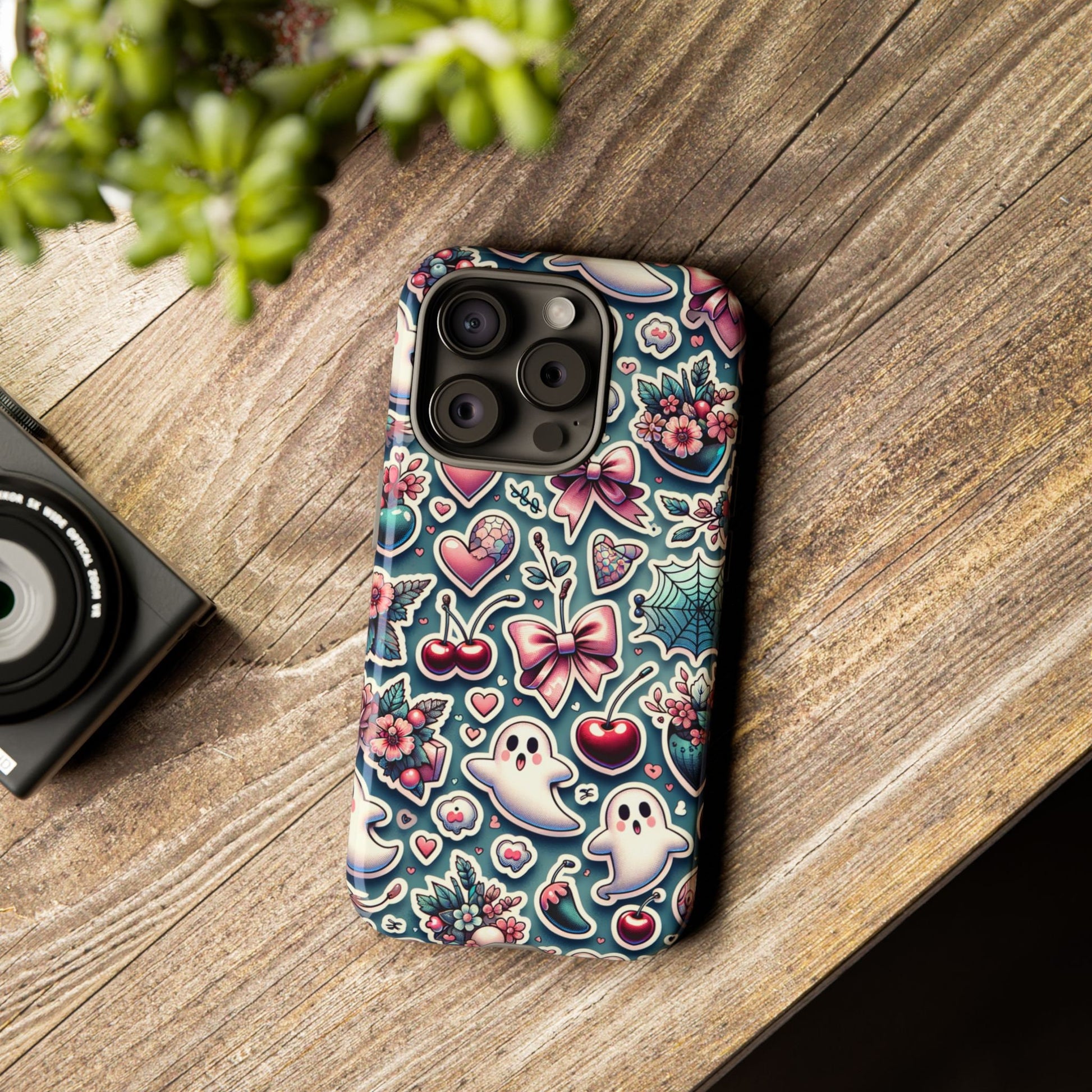 An iPhone with a Cute Coquette Halloween Phone Case featuring a coquette aesthetic design of ghosts, cherries, flowers, and hearts rests on a wooden surface beside a camera and a plant.
