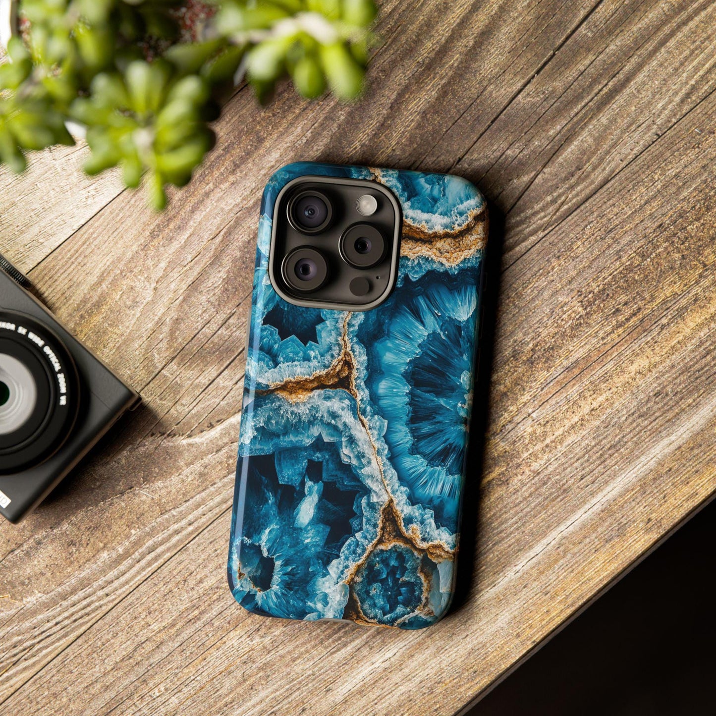 An iPhone sits gracefully on a wooden table, encased in the March Birthstone Aquamarine Crystal Geode Design Phone Case for Aquamarine lovers, while surrounded by a lush plant and an adjacent camera.