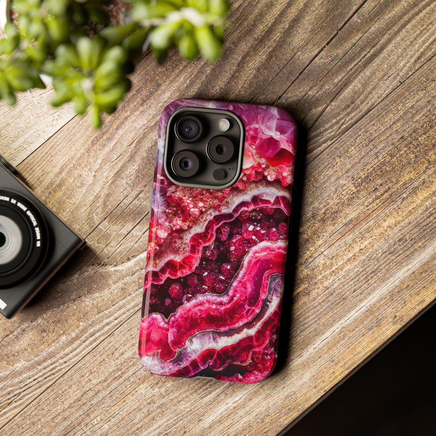 Smartphone with a vibrant, abstract case reminiscent of the July Birthstone Ruby Crystal Geode Design Phone Case, featuring red and pink swirls. It rests gracefully on a wooden surface near a plant and a camera.