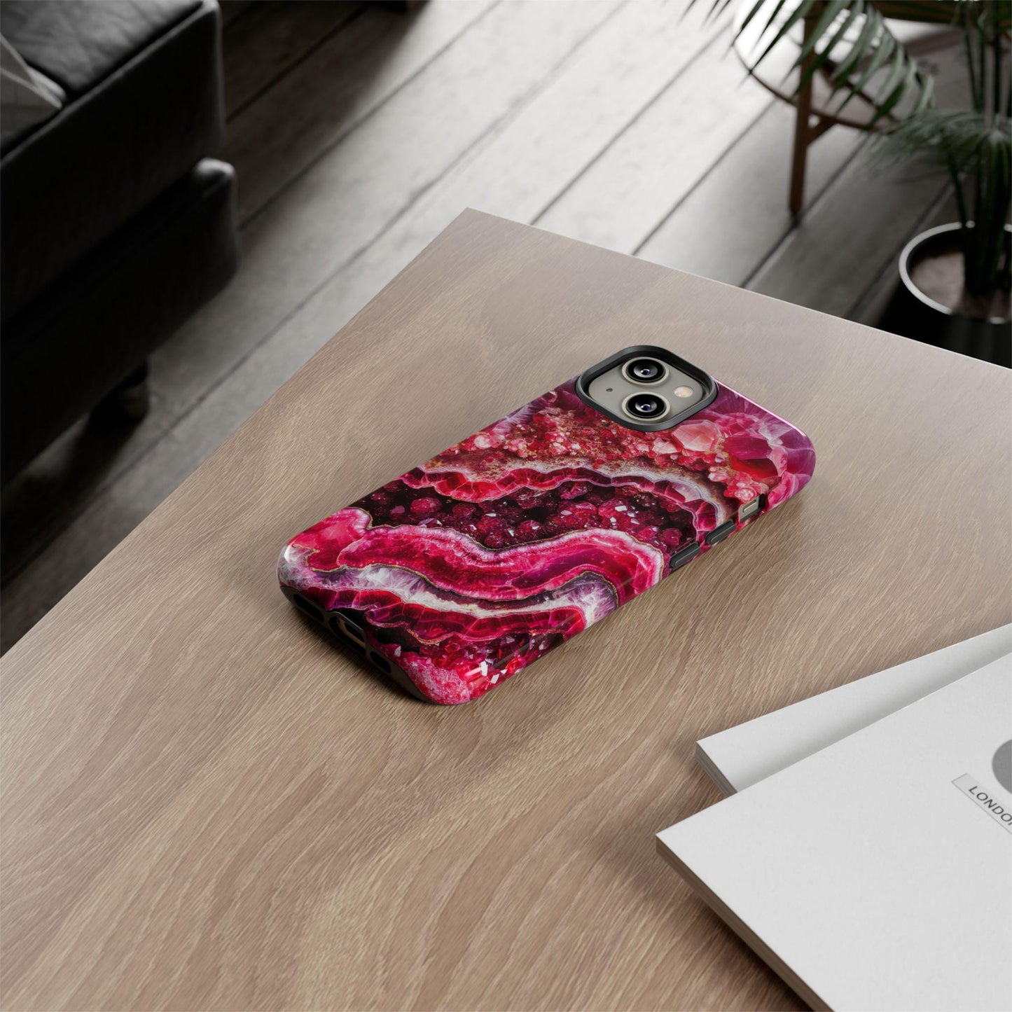 An iPhone featuring the July Birthstone Ruby Crystal Geode Design Phone Case sits on a wooden table beside a stack of papers, with a plant and sofa visible in the background.
