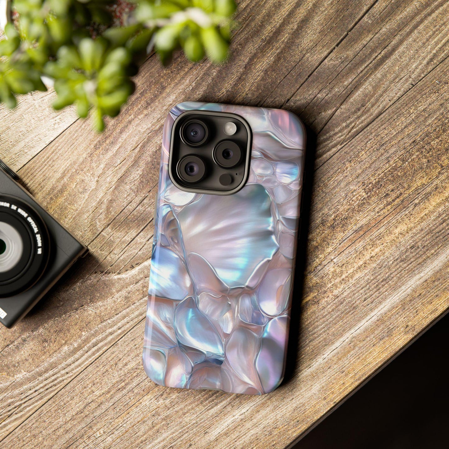 A smartphone encased in a June Birthstone Mother of Pearl Phone Case rests on a wooden surface beside a small plant and a compact camera.
