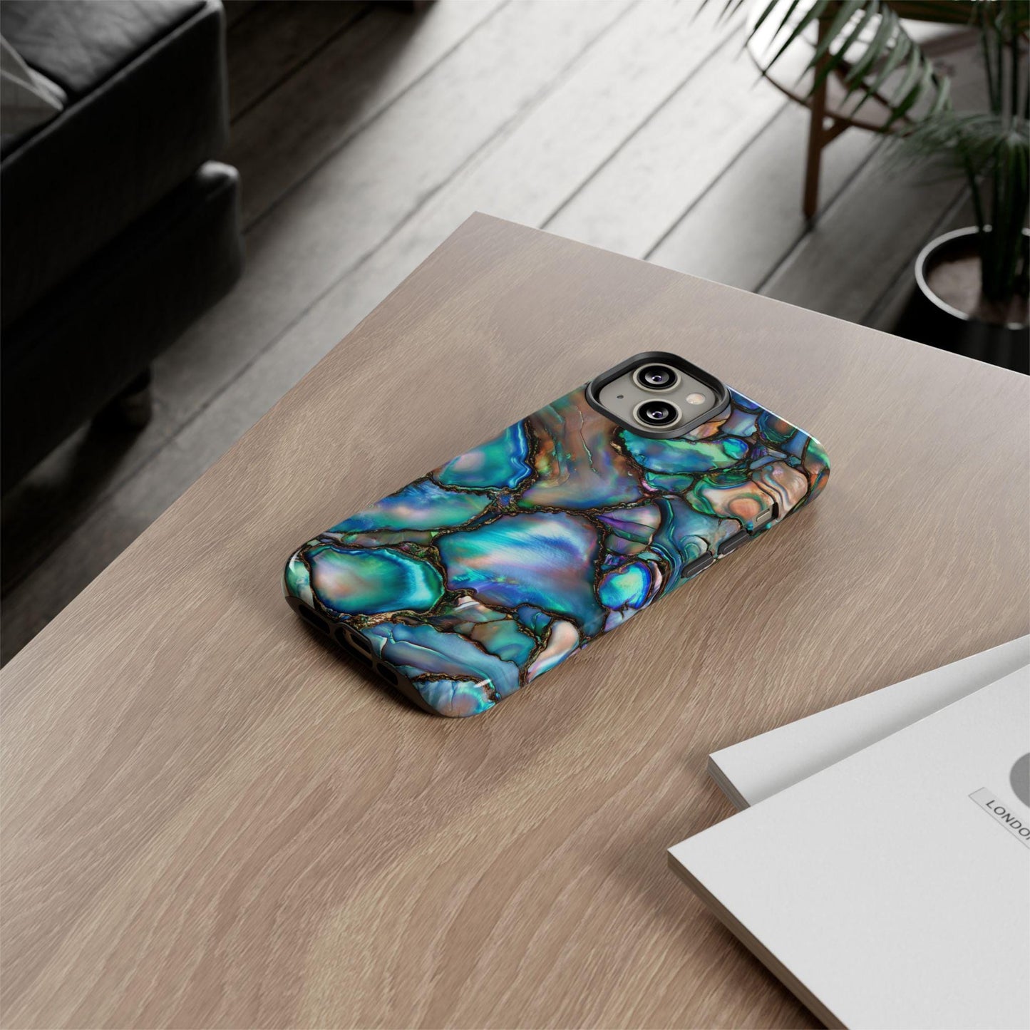 A smartphone with an Abalone Shell Design Tough Phone Casing, featuring Paua Shell Pearlescence, rests on a wooden table next to a white book.