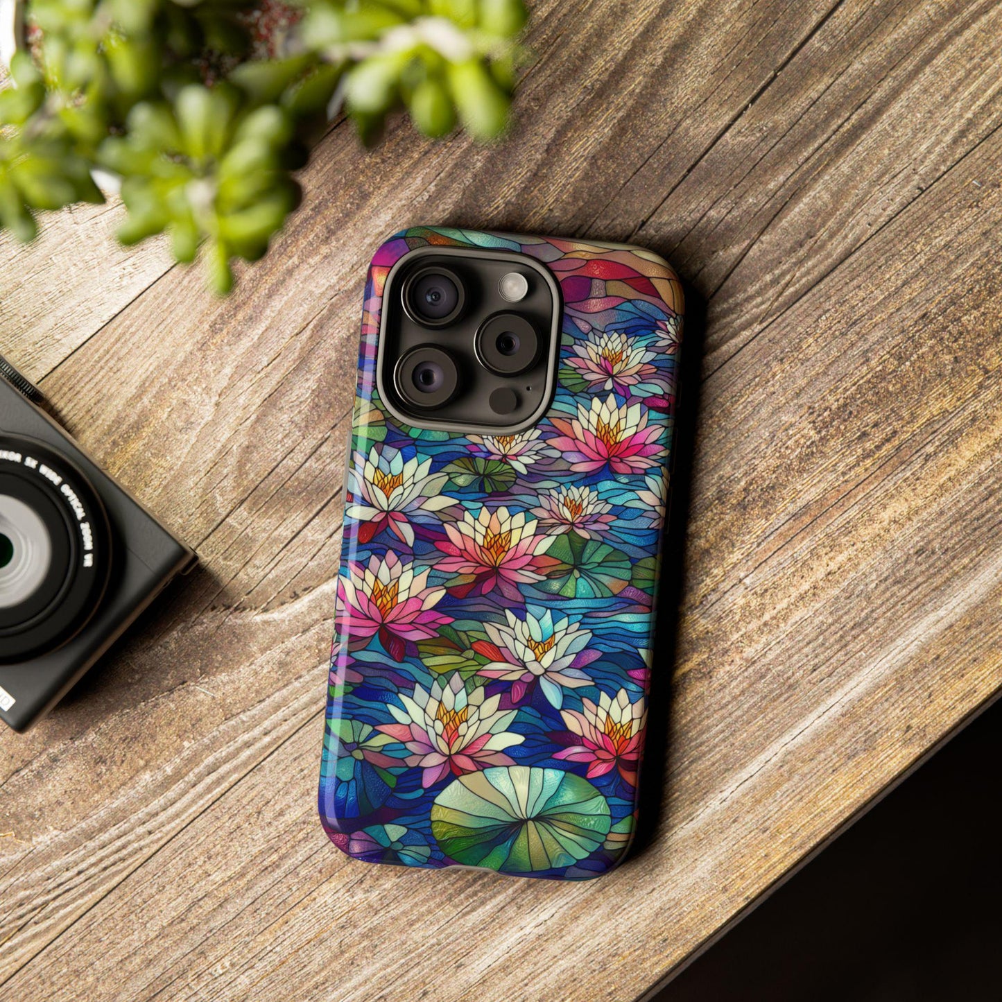A phone adorned with the July Birth Flower Water Lily Wildflower Stained Glass Phone Case rests on a wooden surface beside an elegant camera and a small plant, its design reminiscent of delicate water lilies.