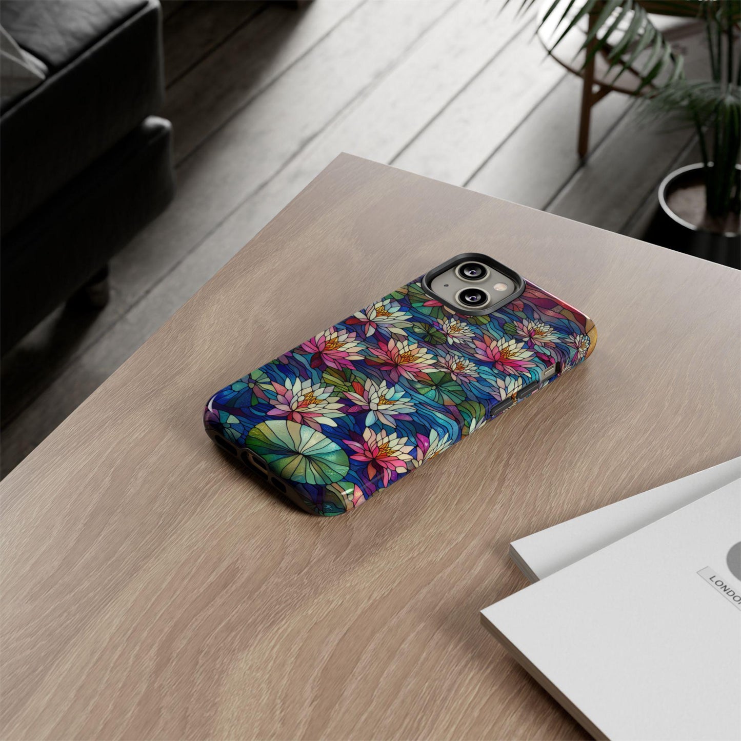 An iPhone with a vibrant "July Birth Flower Water Lily Wildflower Stained Glass" case rests on a wooden table beside a stack of papers.