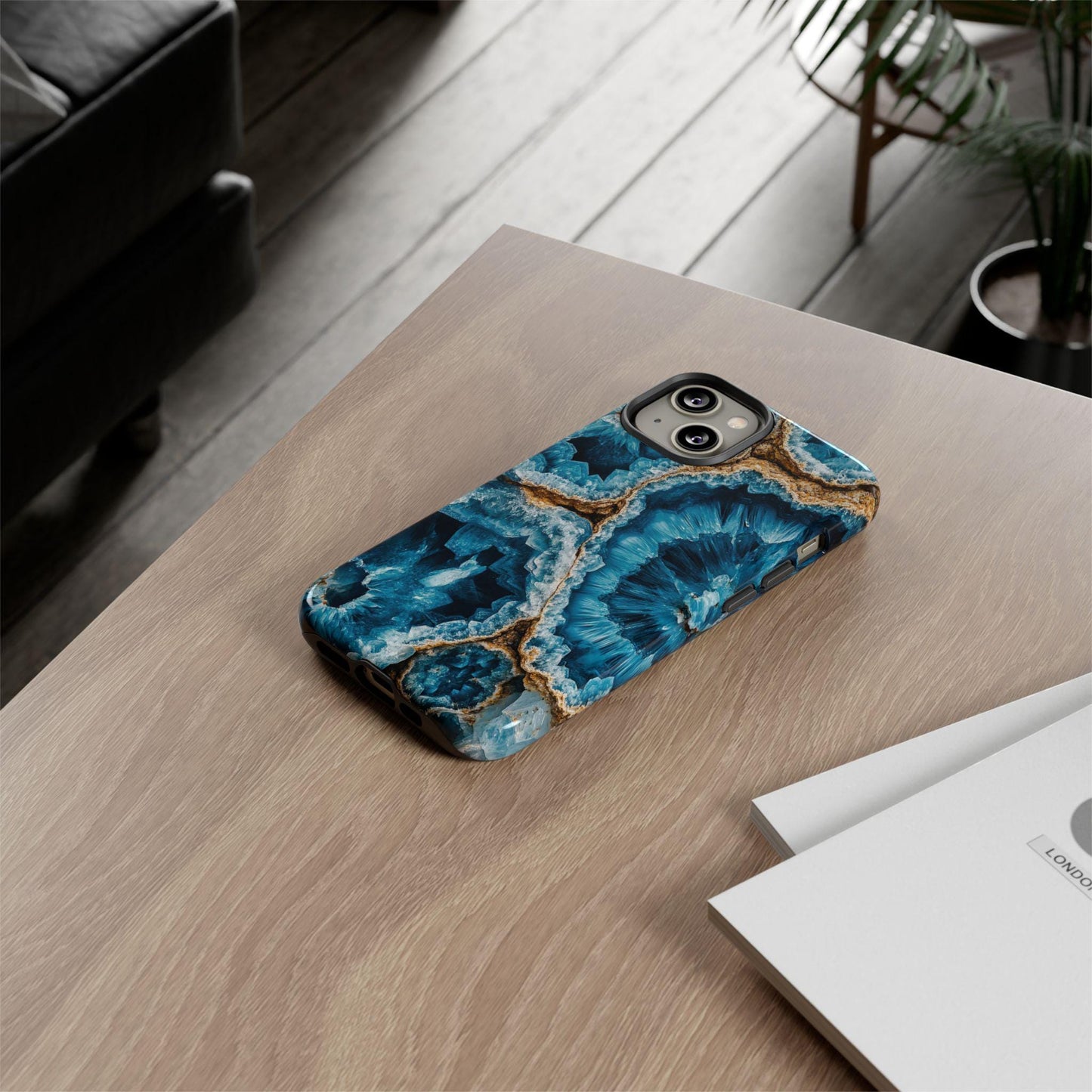 A smartphone equipped with the "March Birthstone Aquamarine Crystal Geode Design Phone Case," perfect for aquamarine lovers, is positioned on a wooden table beside a closed book.