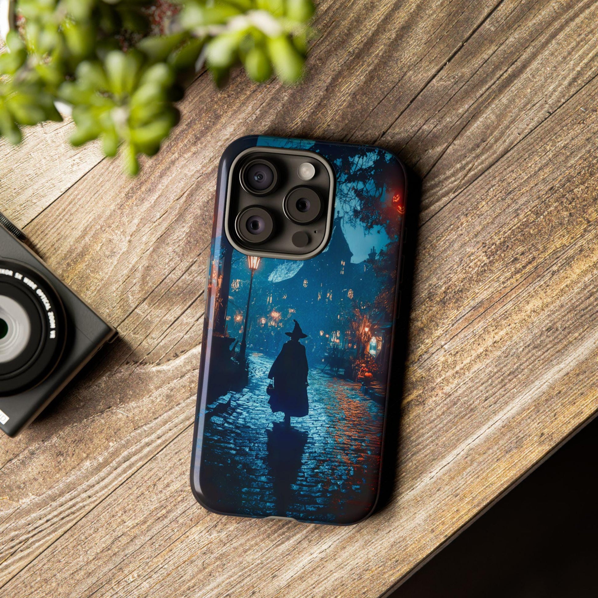 A Witch Walking In Town Halloween Phone Case, featuring dark academia gothic witchy aesthetics, designed for iPhone 15 14 13, Samsung Galaxy, and Google Pixel models, sits on a wooden surface next to a small plant.