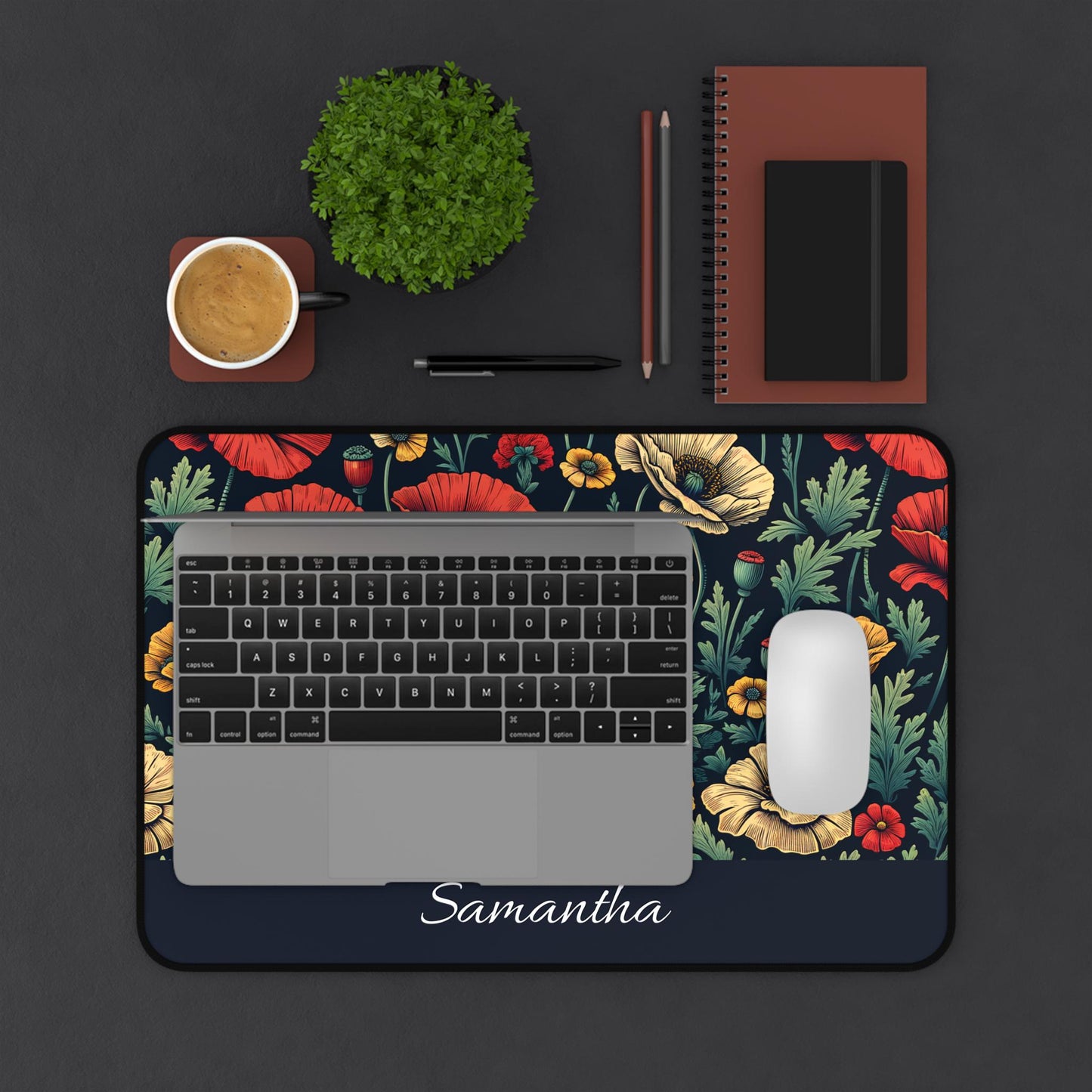 A laptop rests on a Personalized Dark Floral Desk Mat, a Wildflower Poppy Mouse Pad tailored for poppy enthusiasts, adorned with "Samantha." Accompanying it are a white mouse, notebook, two pencils, pen, coffee cup, and plant—all elegantly arrayed on a dark surface.