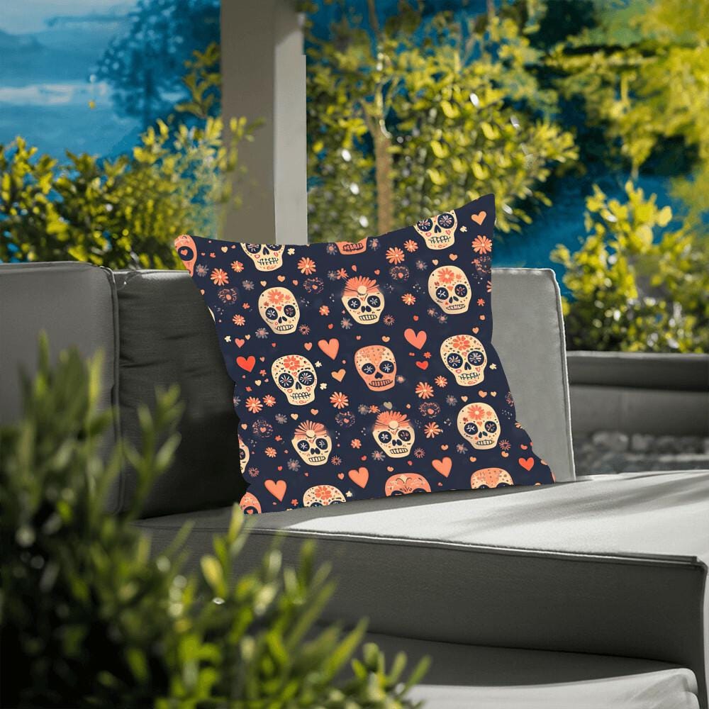 A Day of the Dead Mexican Skull Pillow, featuring a vibrant skull and heart pattern, rests on a gray cushioned outdoor chair, surrounded by lush greenery.