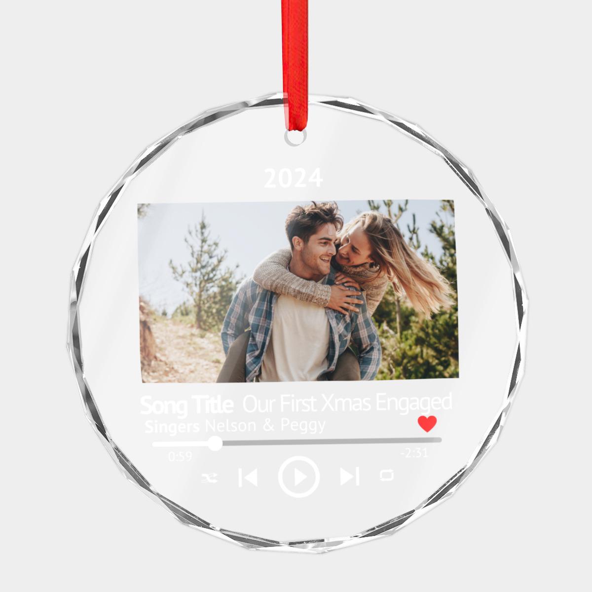 A personalized engagement gift designed to be cherished, this Custom First Christmas Engaged Glass Music Player Photo Ornament showcases a couple outside with the text: "Song Title: Our First Xmas Engaged, Singers: Nelson & Peggy, 2024." Ideal for newly engaged couples to decorate their Xmas tree.