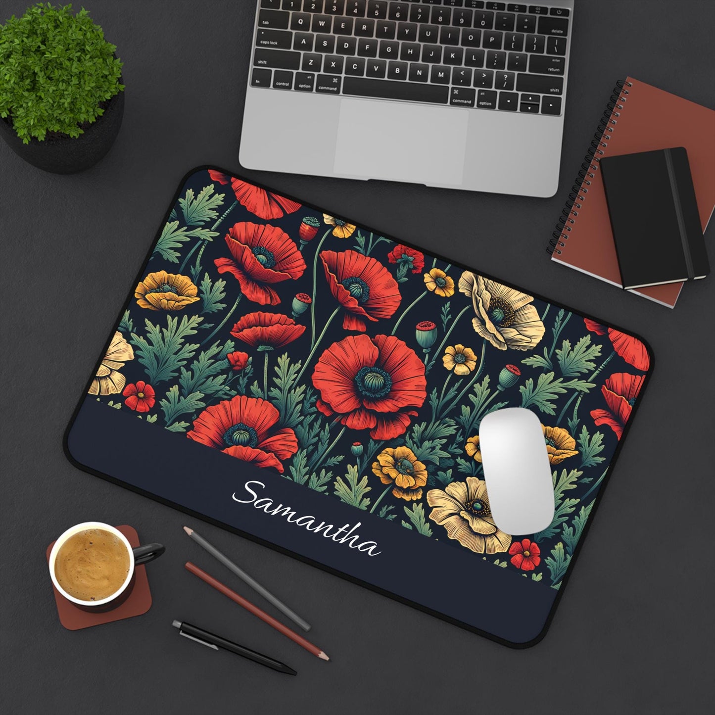 A laptop and notebooks are placed on a dark desk, complemented by a personalized Dark Floral Desk Mat featuring wildflower poppies, with red and yellow blooms alongside the name "Samantha." The inviting workspace is completed with a white mouse, coffee cup, pen, and potted plant.