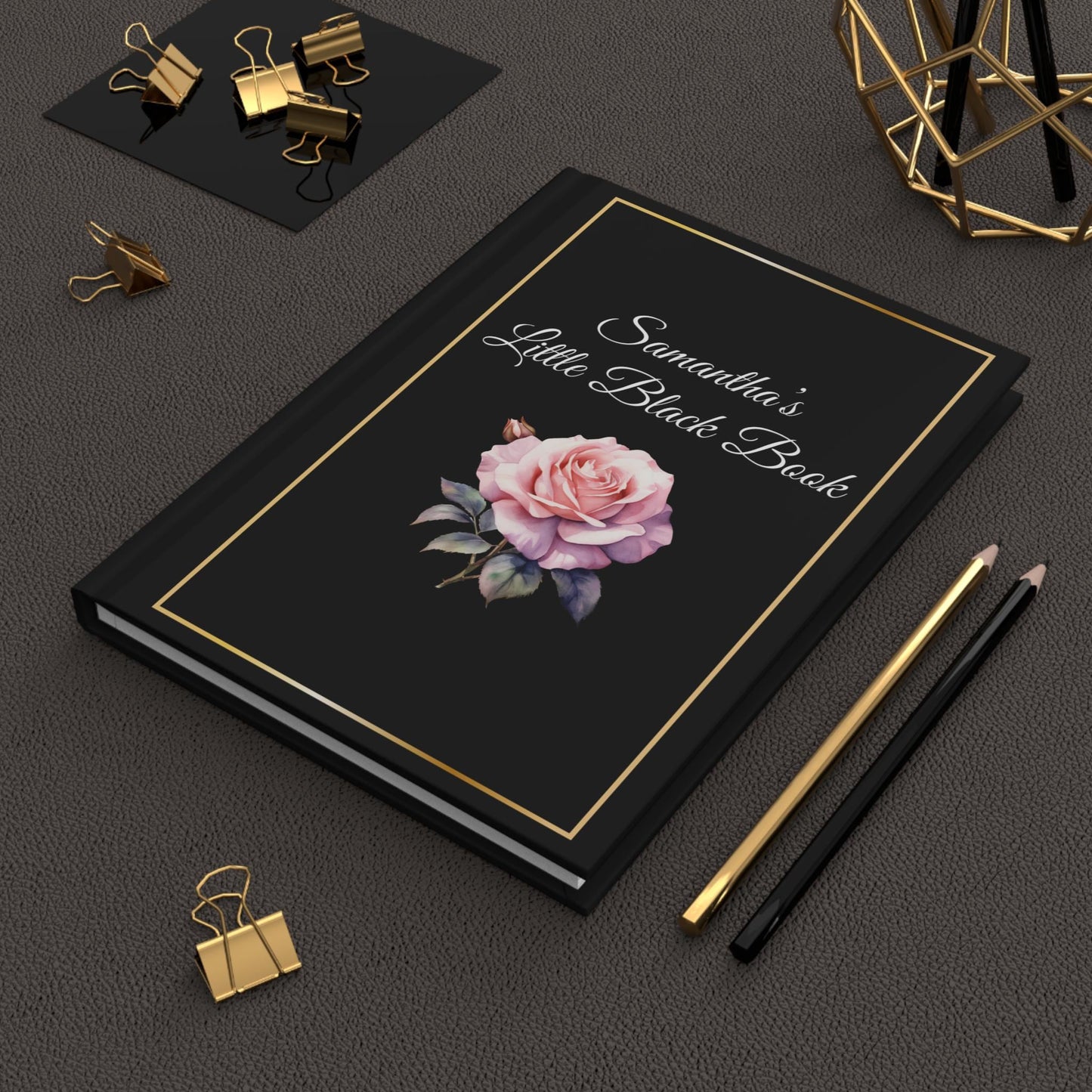 A custom notebook featuring "Samantha's Little Black Book" and adorned with a rose on the cover, comes with a personalized birth flower theme. This elegant journal set includes a pen, pencils, gold clips, and decorative items laid out on a textured surface; an ideal gift for your daughter to stylishly capture her thoughts.