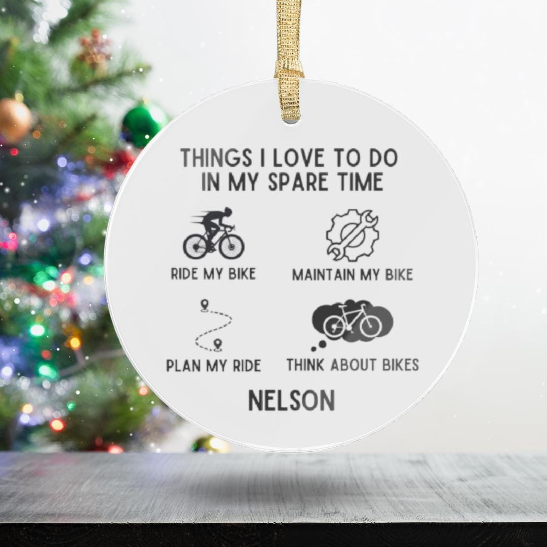 The Personalized Cyclist Ornament, crafted from transparent acrylic, showcases "Things I love to do in my spare time" with icons representing riding, maintaining, and planning bike activities. Elegantly customized with the name "Nelson" at the bottom, this ornament makes an ideal gift for cycling enthusiasts. It complements a beautifully adorned Christmas tree and sets a festive atmosphere.