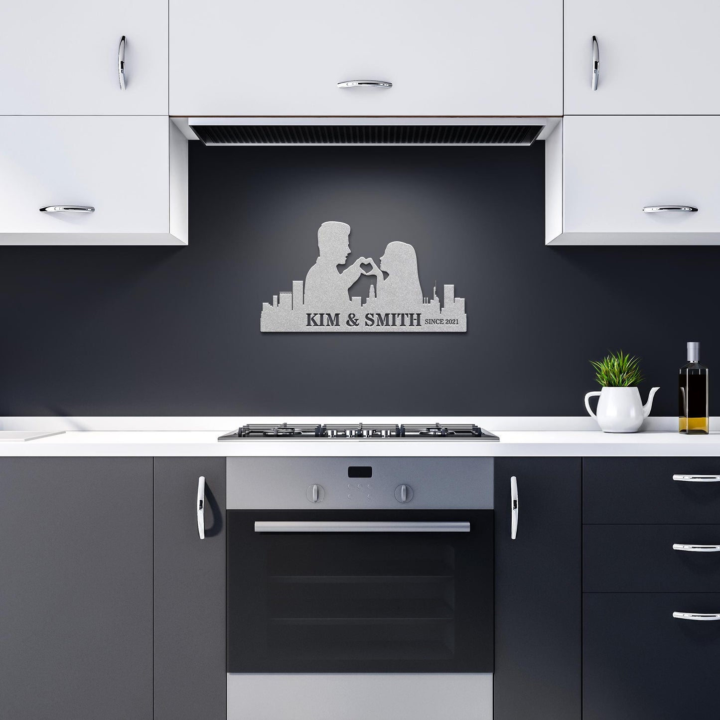 Modern kitchen with white and gray cabinets, an oven, and a Personalized USA City Skyline Couple Metal Sign featuring silhouettes and the text "Kim & Smith Since 2021." A small potted plant and an oil bottle are on the counter.