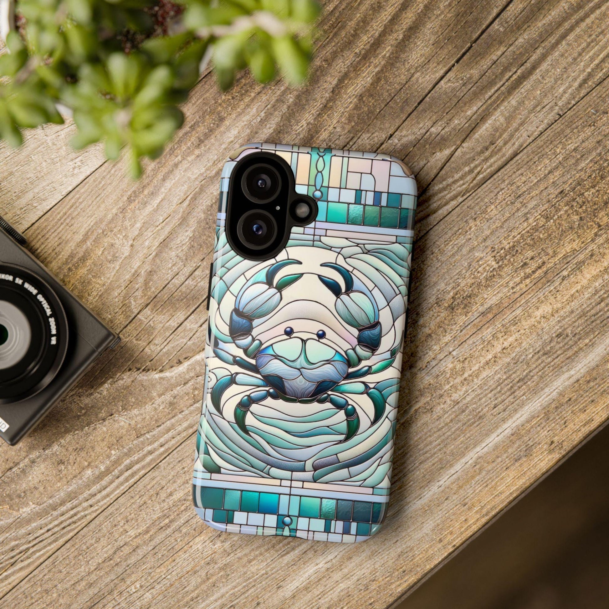 A smartphone with a Zodiac Cancer Faux Stained Glass Mosaic Phone Case, featuring an astrology celestial horoscope design, rests on a wooden table beside a camera and plant.