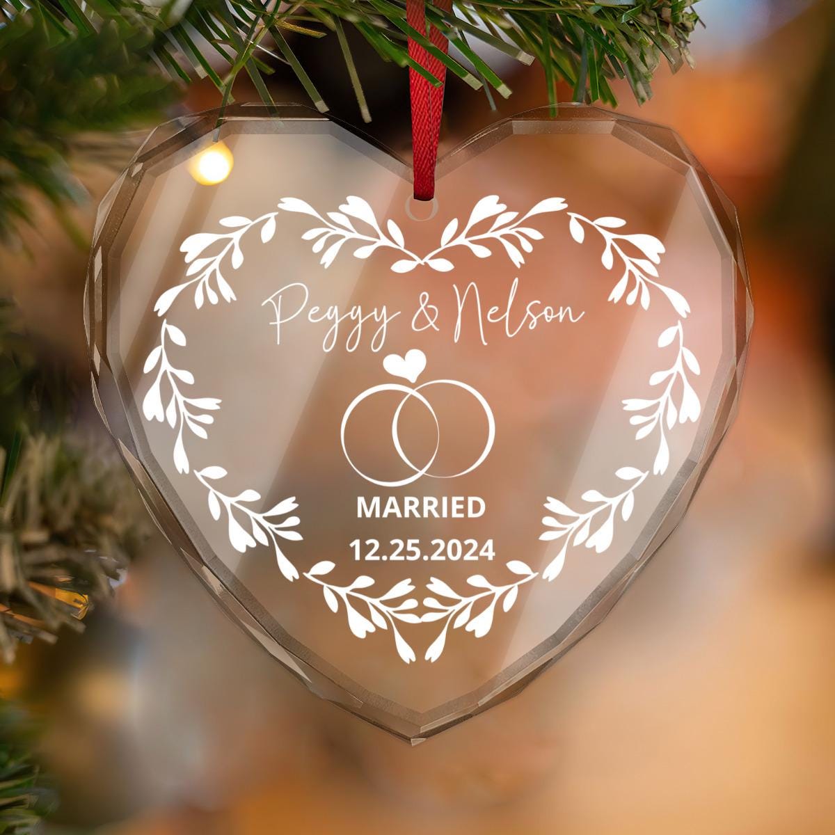A heart-shaped, crystal glass ornament with a floral design, featuring intertwined rings and the text: "Peggy & Nelson Married 12.25.2024," hangs elegantly on a Christmas tree—perfect as a personalized keepsake for married couples' festive celebrations.