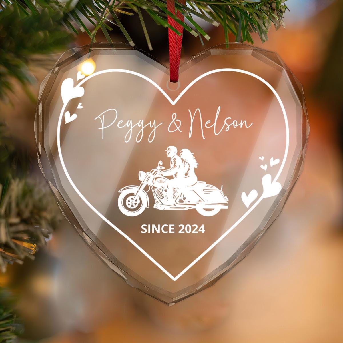 A custom heart-shaped glass ornament with a couple design and the labels "Peggy & Nelson" and "SINCE 2024" hangs from a red ribbon. Ideal as a biker couple keepsake or gift.