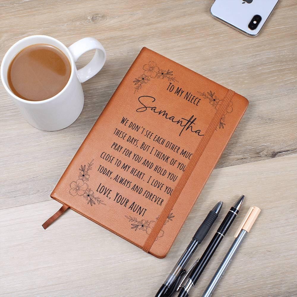 A Personalized Leather Journal For Niece, inscribed with "Samantha" from her aunt, sits on a table with coffee, a smartphone, and three pens—an ideal teenage girl gift.