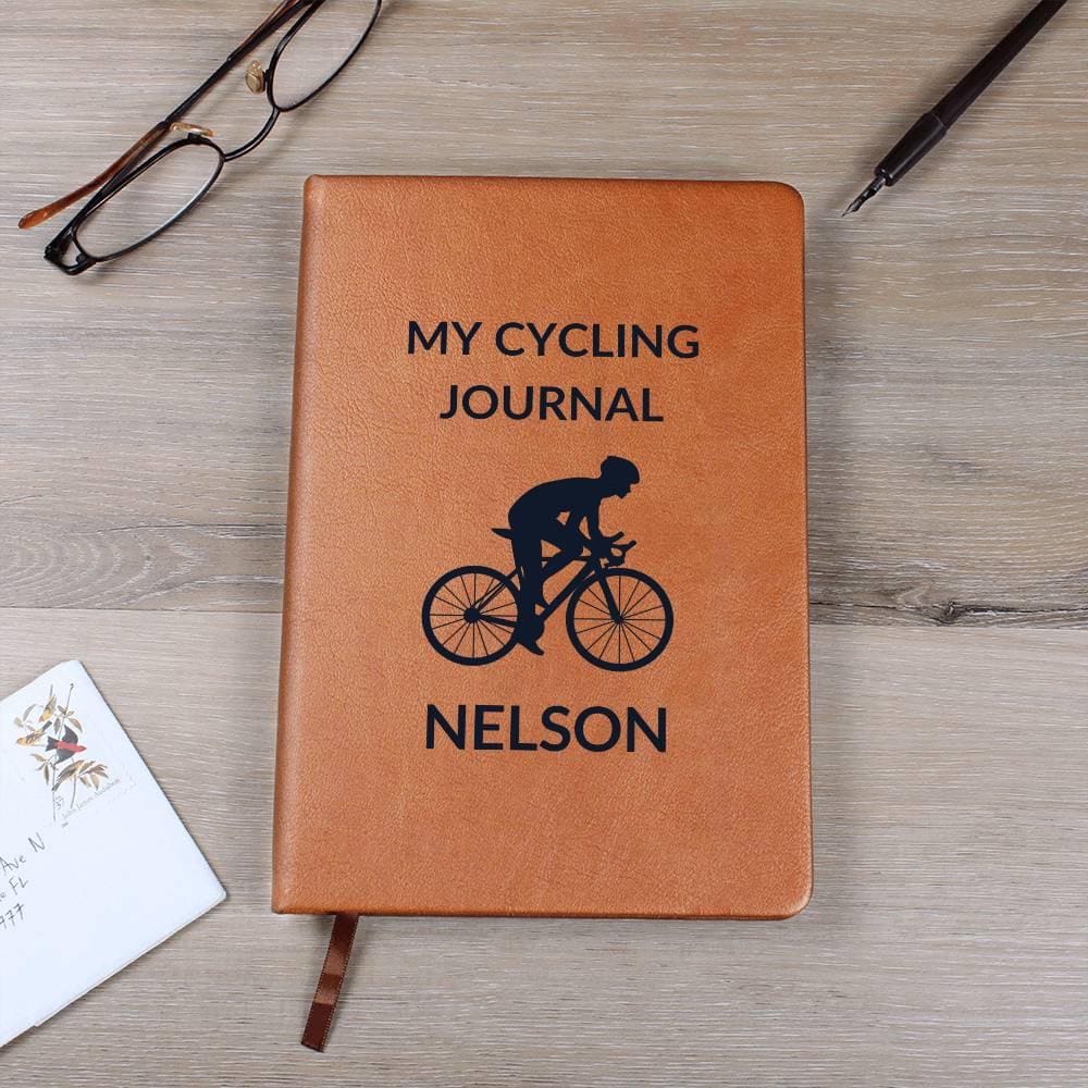 The brown "My Cycling Journal," personalized leather cycling journal features a cyclist graphic and rests on wood with glasses, pen, and paper; perfect for jotting adventures in your log book. Customize this notebook with a ribbon bookmark—ideal for road bikers, bike lovers & cyclists.