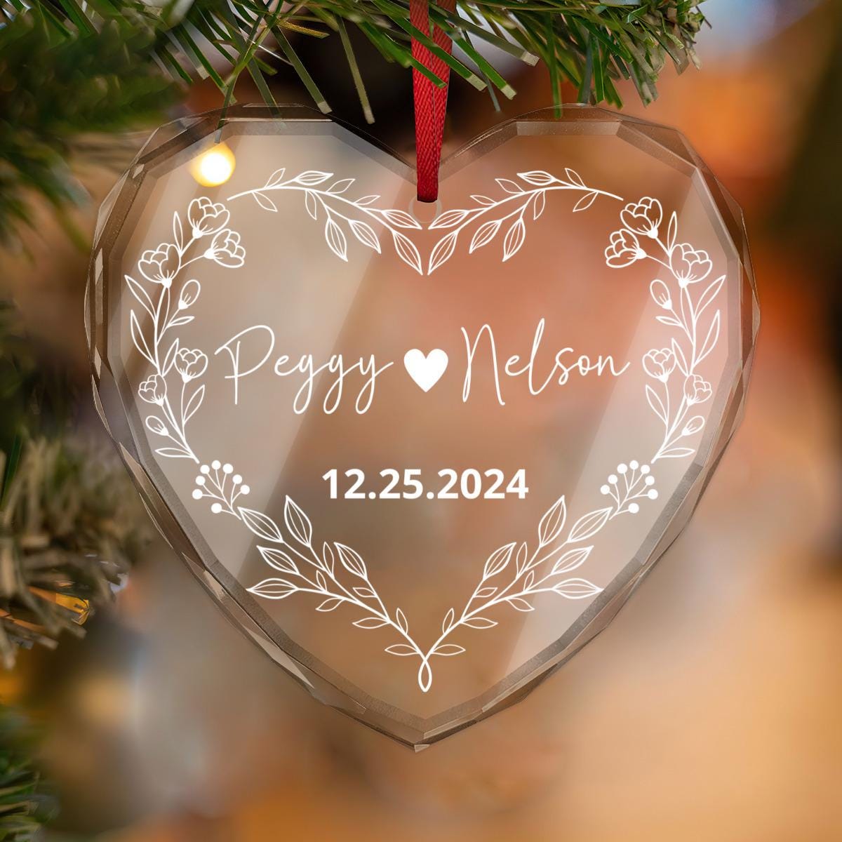 A heart-shaped Custom Couple CRYSTAL GLASS Ornament featuring floral designs hangs on a tree with "Peggy Nelson" and "12.25.2024," making it the perfect personalized gift and keepsake for couples to cherish forever.