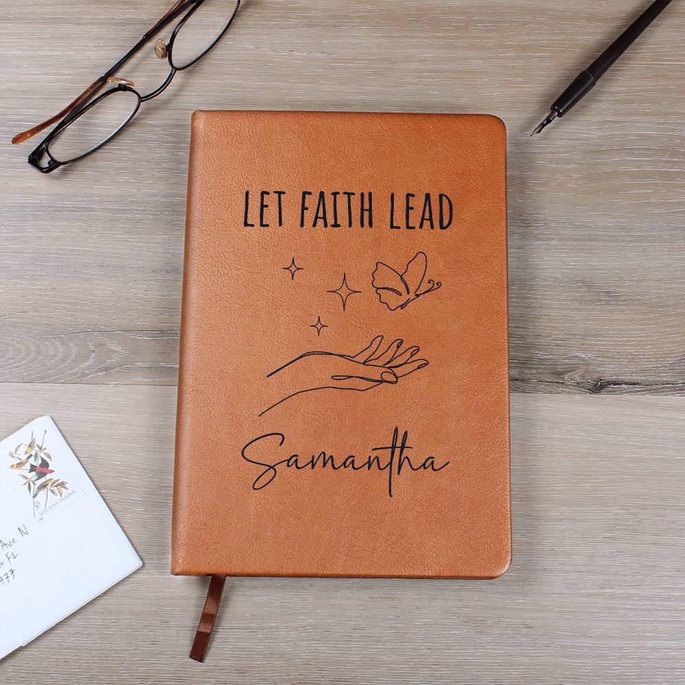 The Personalized Leather Prayer Journal features "Let Faith Lead" and "Samantha" on a brown cover with a hand, stars, and butterfly design. It's surrounded by glasses, a pen, and an envelope on wood. This custom devotional notebook makes cherished religious gifts.