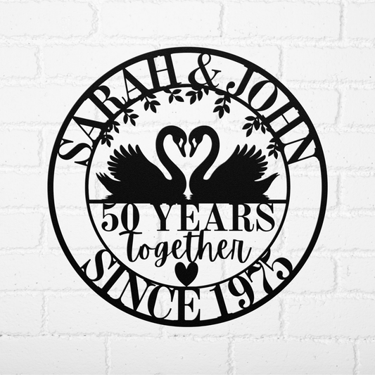 Personalized 50th Anniversary Metal Wall Art: Black circular sign with two swans forming a heart, decorative leaves and hearts, and custom text—"SARAH & JOHN, 50 YEARS together, SINCE 1973"—displayed on a white brick wall.