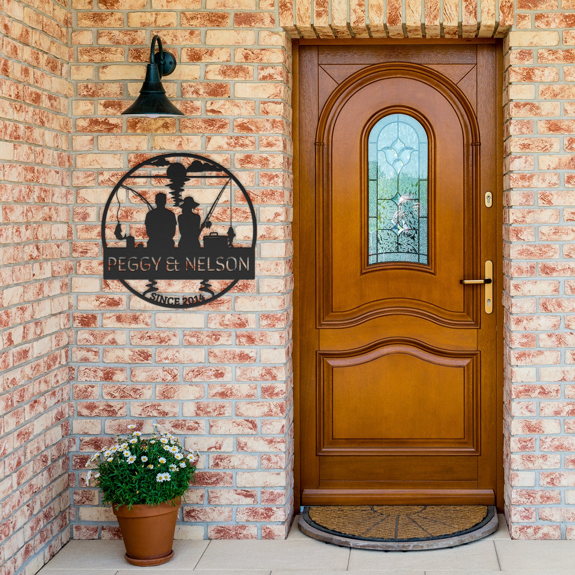 A wooden door with a stained glass window and a metal sign reading "Peggy & Nelson, Since 2015" is mounted on a brick wall, with the Couple Fishing Together Metal Wall Art displayed beside it. A potted plant with white flowers sits near the door.