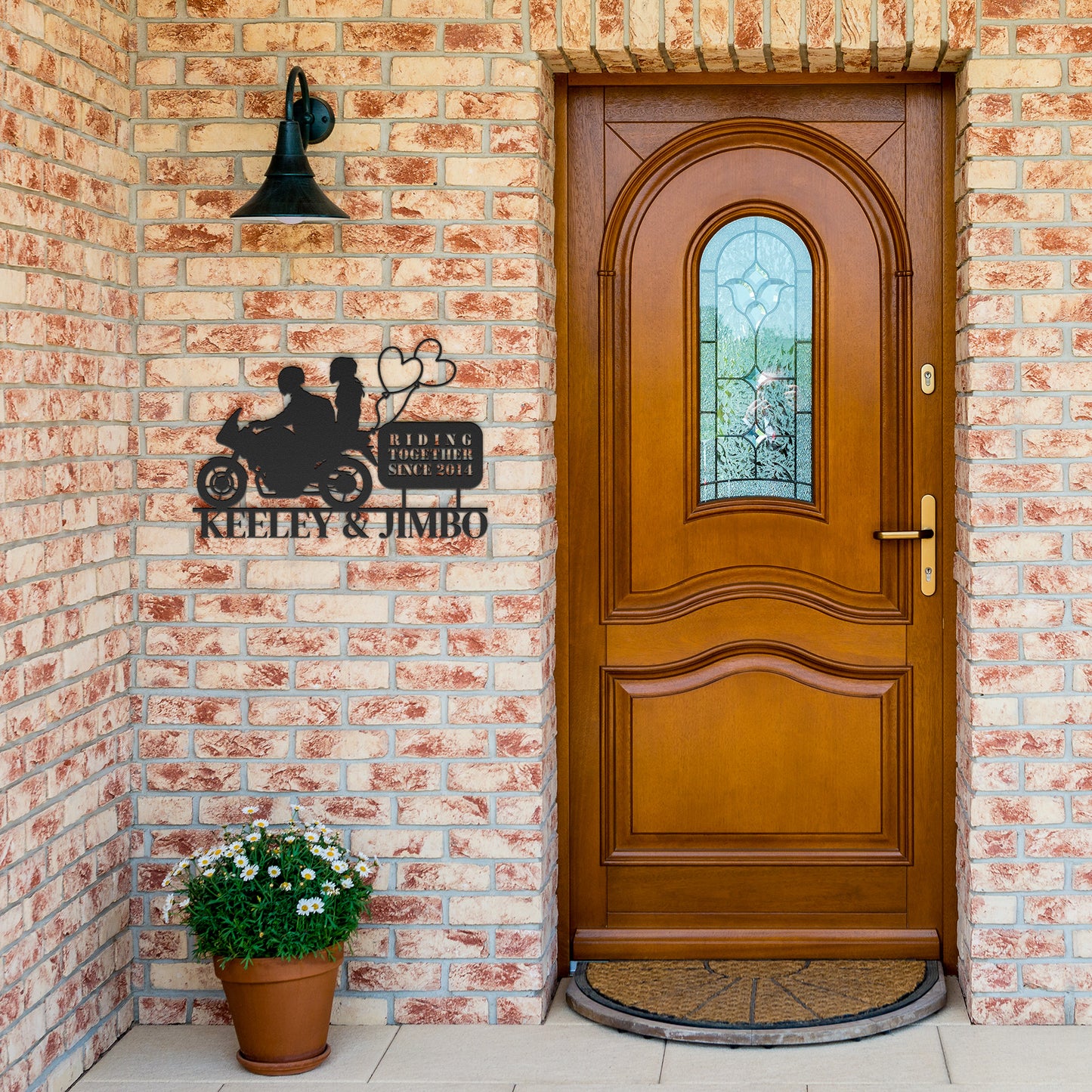 A wooden front door with a stained glass window, next to a brick wall with Custom Motorcycle Couple Metal Wall Art and a potted plant.