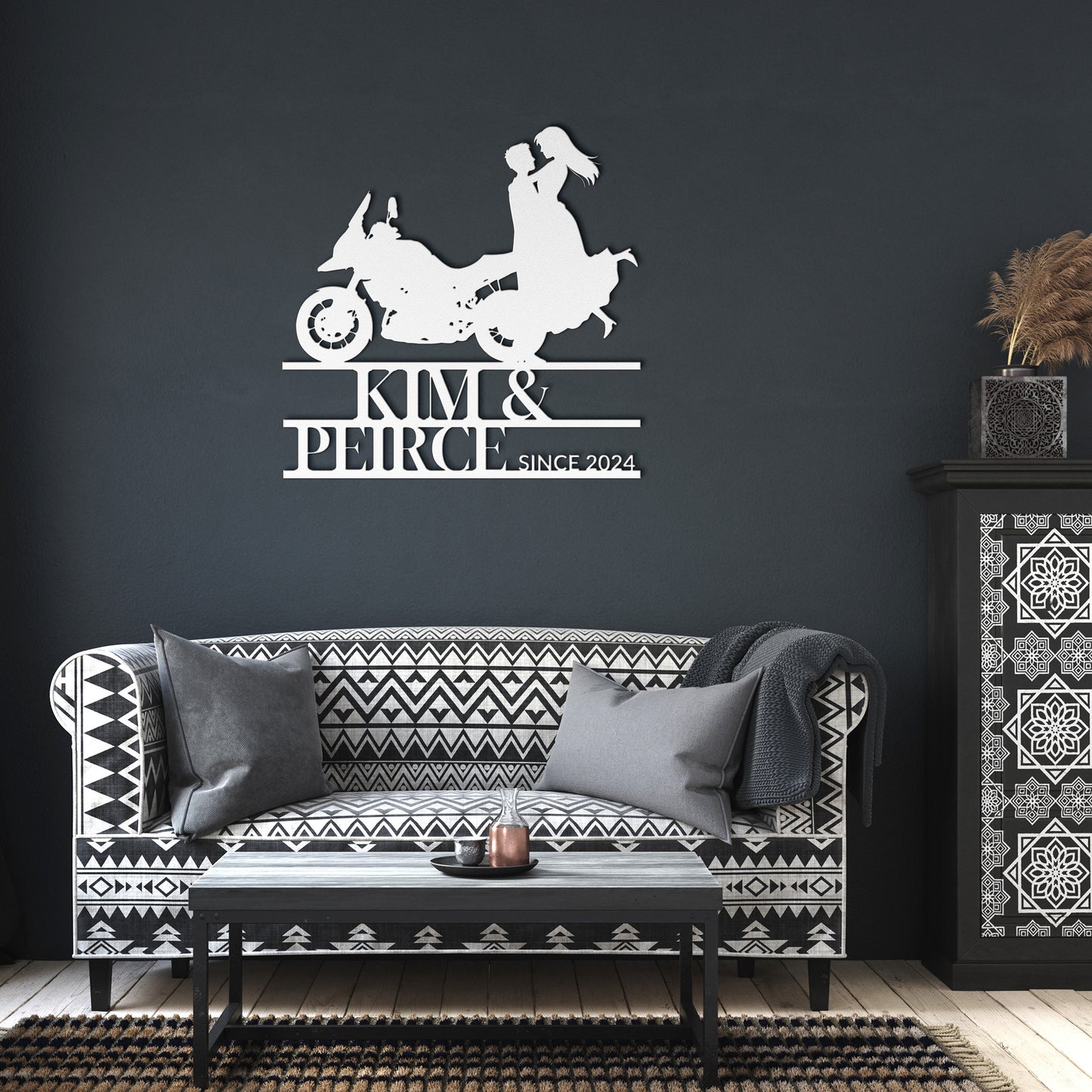 The Custom Motorcycle Couple Metal Wall Art, made from 18 gauge steel, features a couple on a motorcycle with the text "Kim & Peirce Since 2024," bringing a distinctive flair to your home decor. It is stylishly positioned above a patterned sofa with pillows and paired with a decorative cabinet.
