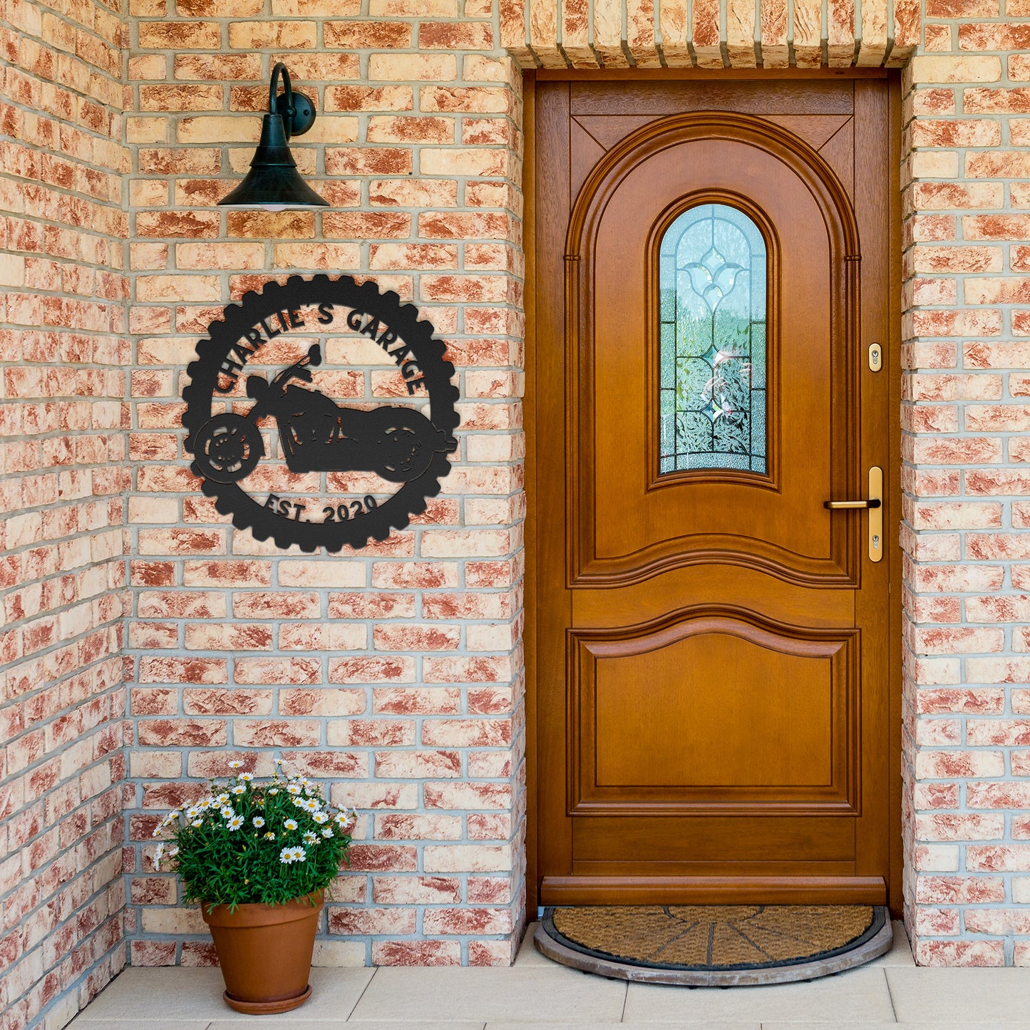 A wooden front door set in a brick wall with a stained glass window. A Custom Motorcycle Garage Sign reading "Charlie's Garage Est. 2021" and a black light are mounted to the wall, enhancing the home decor. A potted plant sits beside the door.