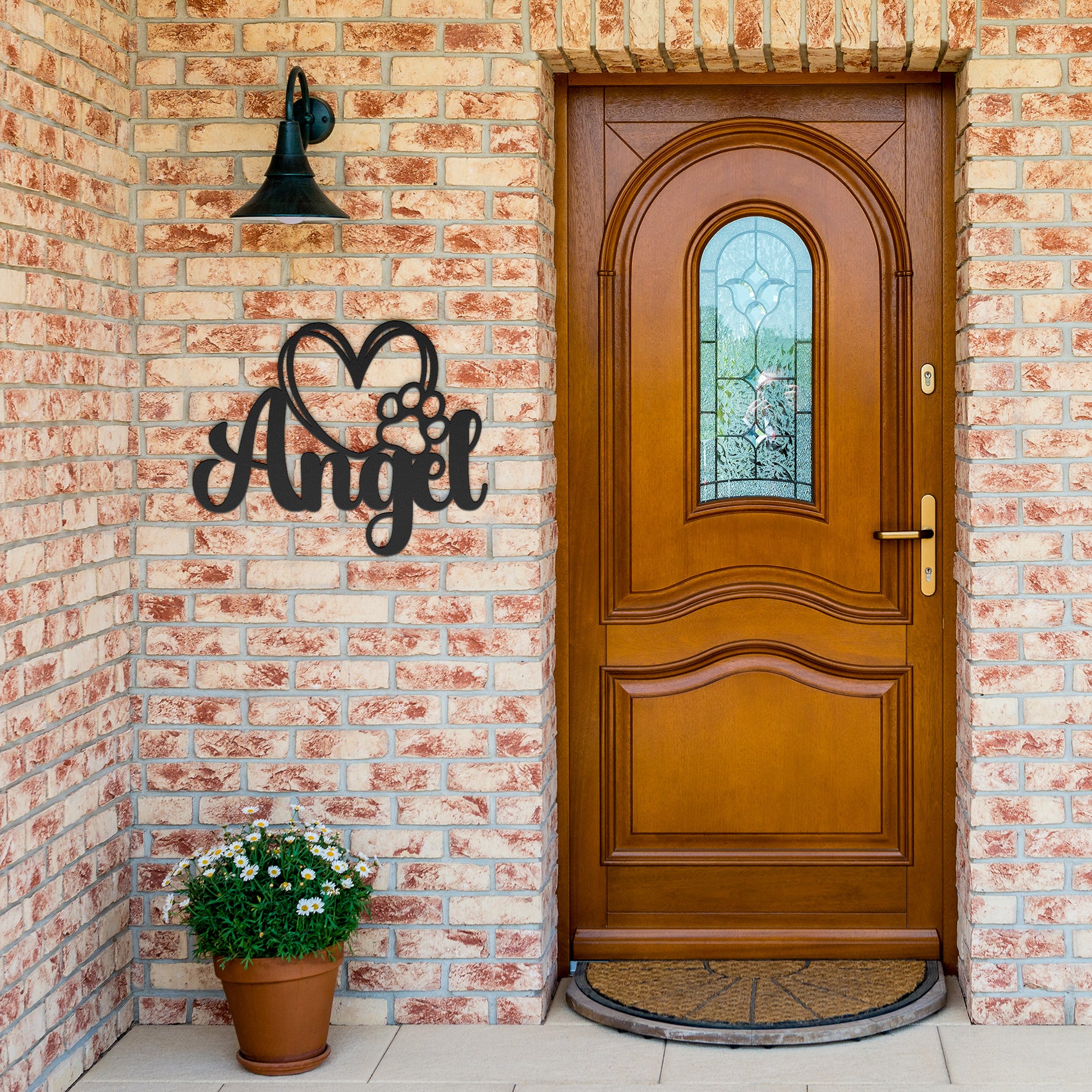 A wooden door with a glass panel in the center is set in a brick wall. To the left of the door is a potted plant with white flowers, and an 18 gauge steel sign that reads "Angel" with a heart and pawprint adds charm to the home decor. This unique touch is provided by the Custom Pet Name Metal Sign with Heart and Paw.