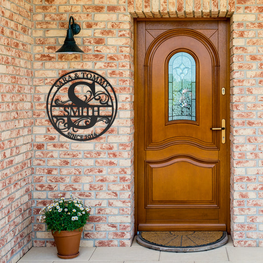 A wooden front door set in a brick wall with a Custom Round Personalized Metal Monogram Sign on the left and a potted plant at the bottom, adding a unique touch to your home decor.