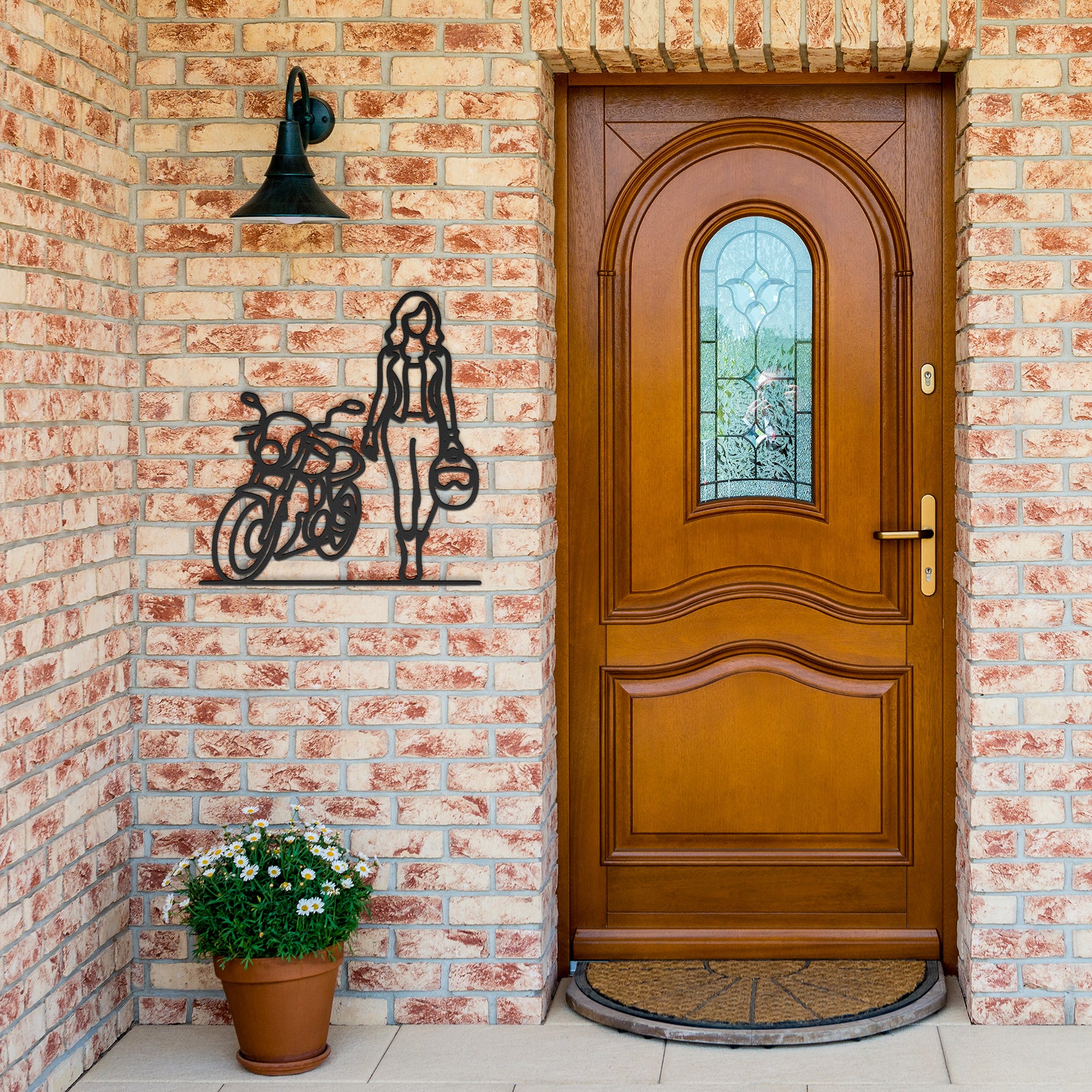 A wooden door with a decorative glass panel stands beside a FEMALE BIKER LINE ART mounted on the brick wall, complemented by a black wall lantern above and a potted plant with white flowers on the patio—creating charming home decor that could be accented further with a tasteful metal sign.