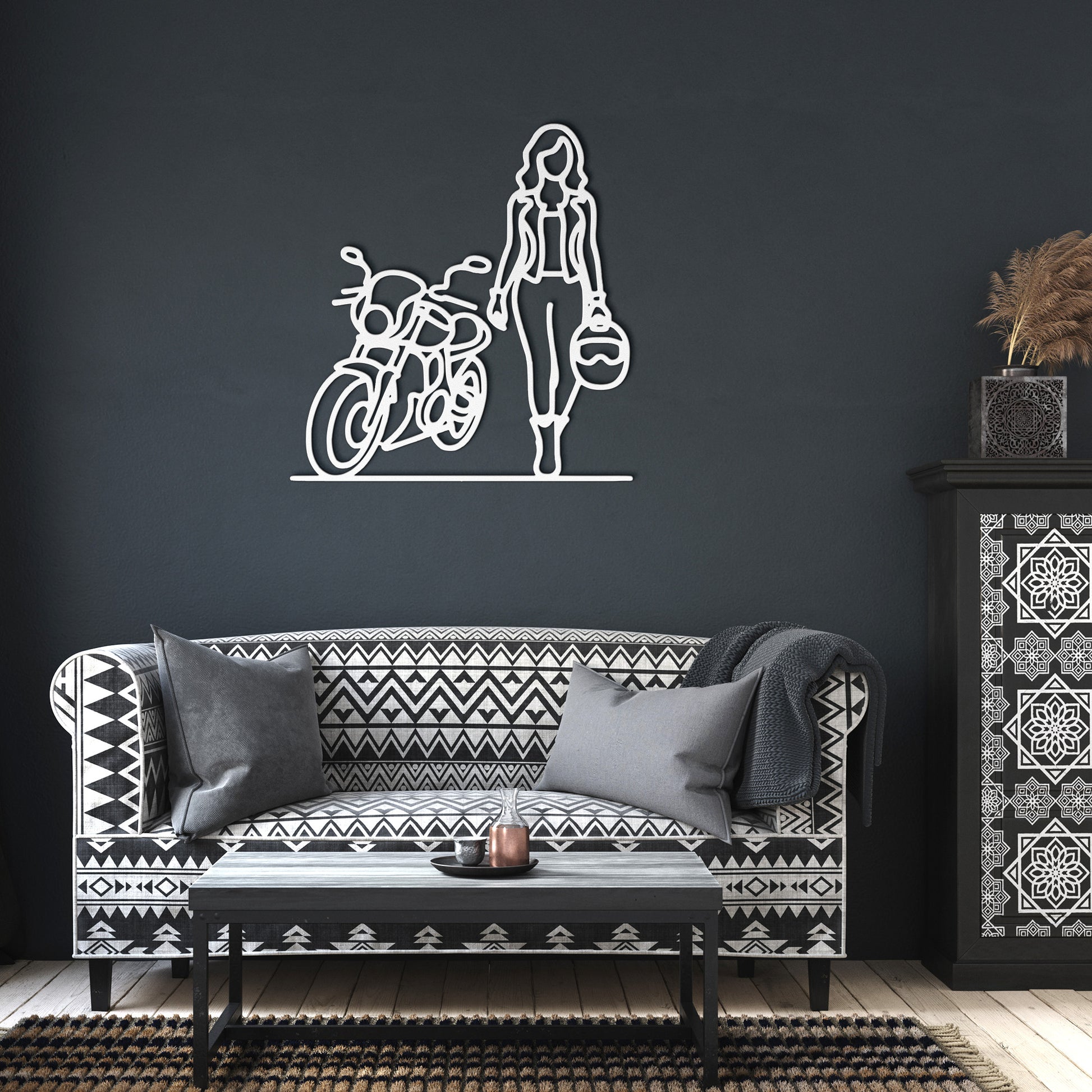 A powder-coated sign featuring the FEMALE BIKER LINE ART is elegantly displayed above a patterned sofa adorned with cushions. To the side, a decorated cabinet with plants enhances the charm of the home decor.