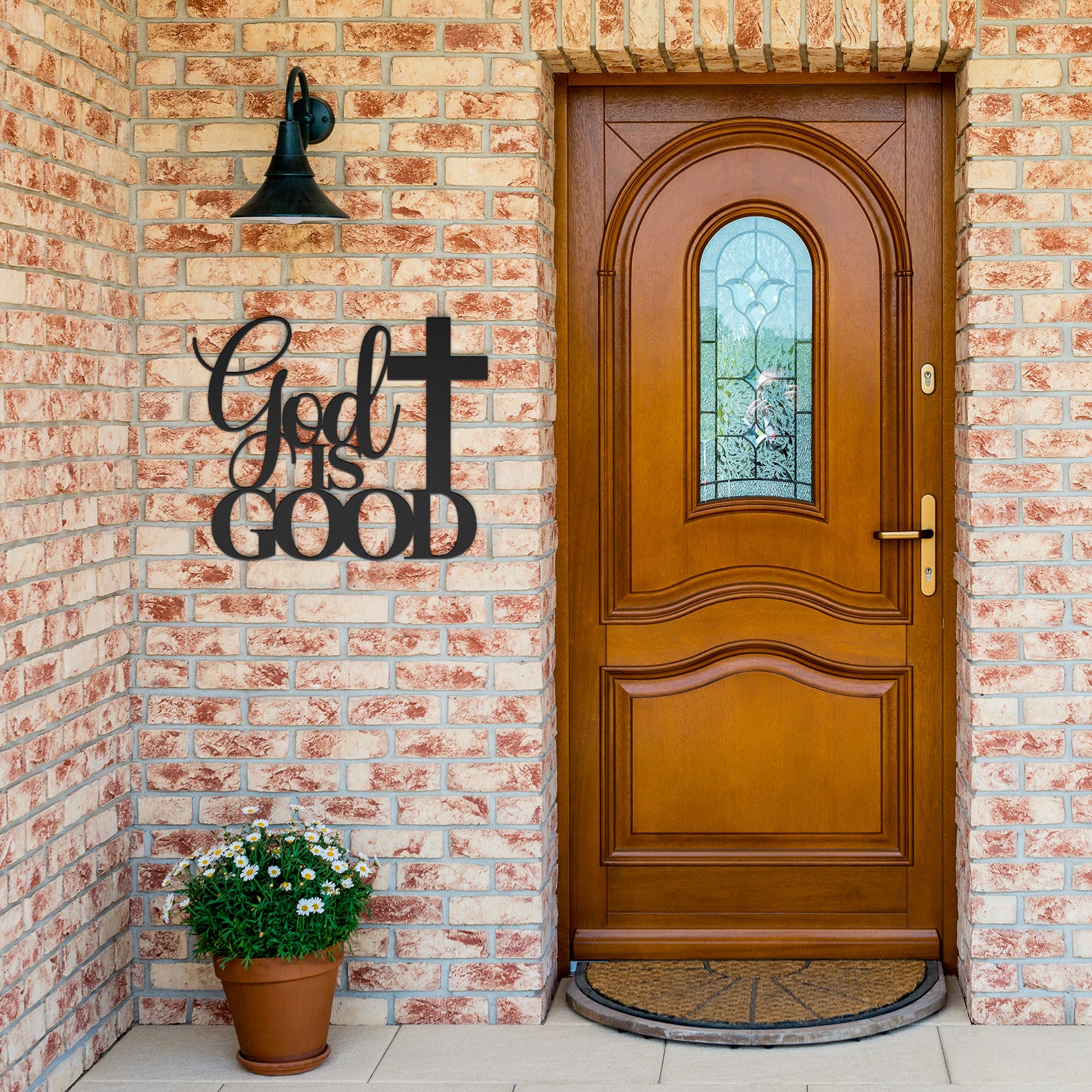 A brown wooden door with decorative glass is set in a brick wall. A potted plant sits beside it, and a stylish powder-coated sign "God Is Good Christian Metal Art" next to a cross enhances the decor. A black outdoor lamp hangs above the sign, completing the charming home decor scene.